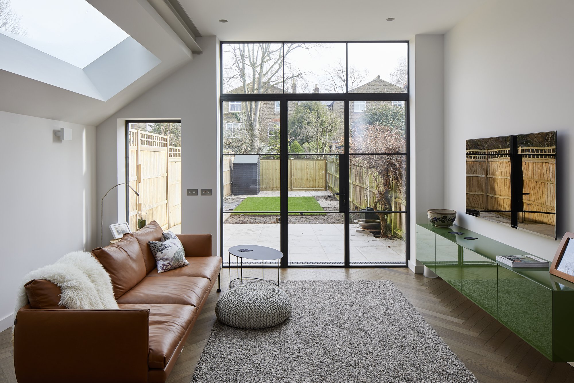 A modern living room with a brown leather sofa and a grey pouf on a textured rug. Large glass french doors open to a patio with a small garden. A green cabinet holds a TV, and a skylight brightens the space. Minimalist decor creates a cozy atmosphere