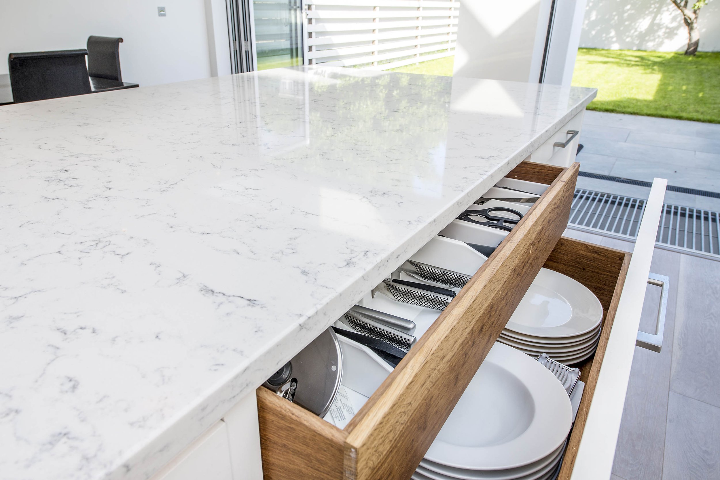A modern kitchen featuring a marble countertop with an open drawer showcasing neatly arranged plates and cutlery. Natural light enters through large windows, reflecting off the hardwood floors and high-end appliances, highlighting the clean, contempo