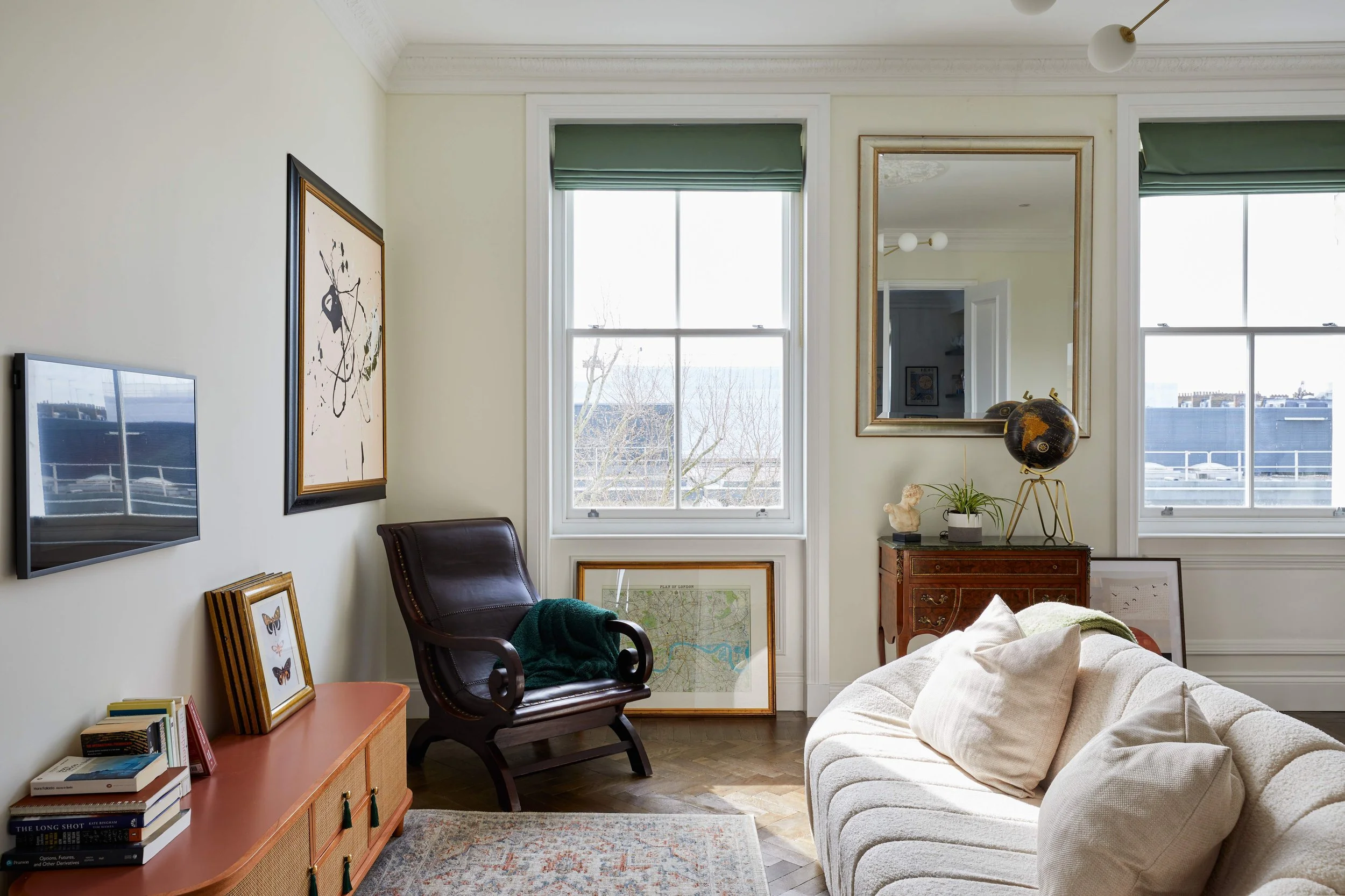 A cozy living and dining room in a Georgian townhouse with a white sofa, decorative pillows, and a dark wooden table with chairs. Large windows fill this Pimlico gem with natural light, while an ornate mirror hangs above a vintage cabinet. Artworks a