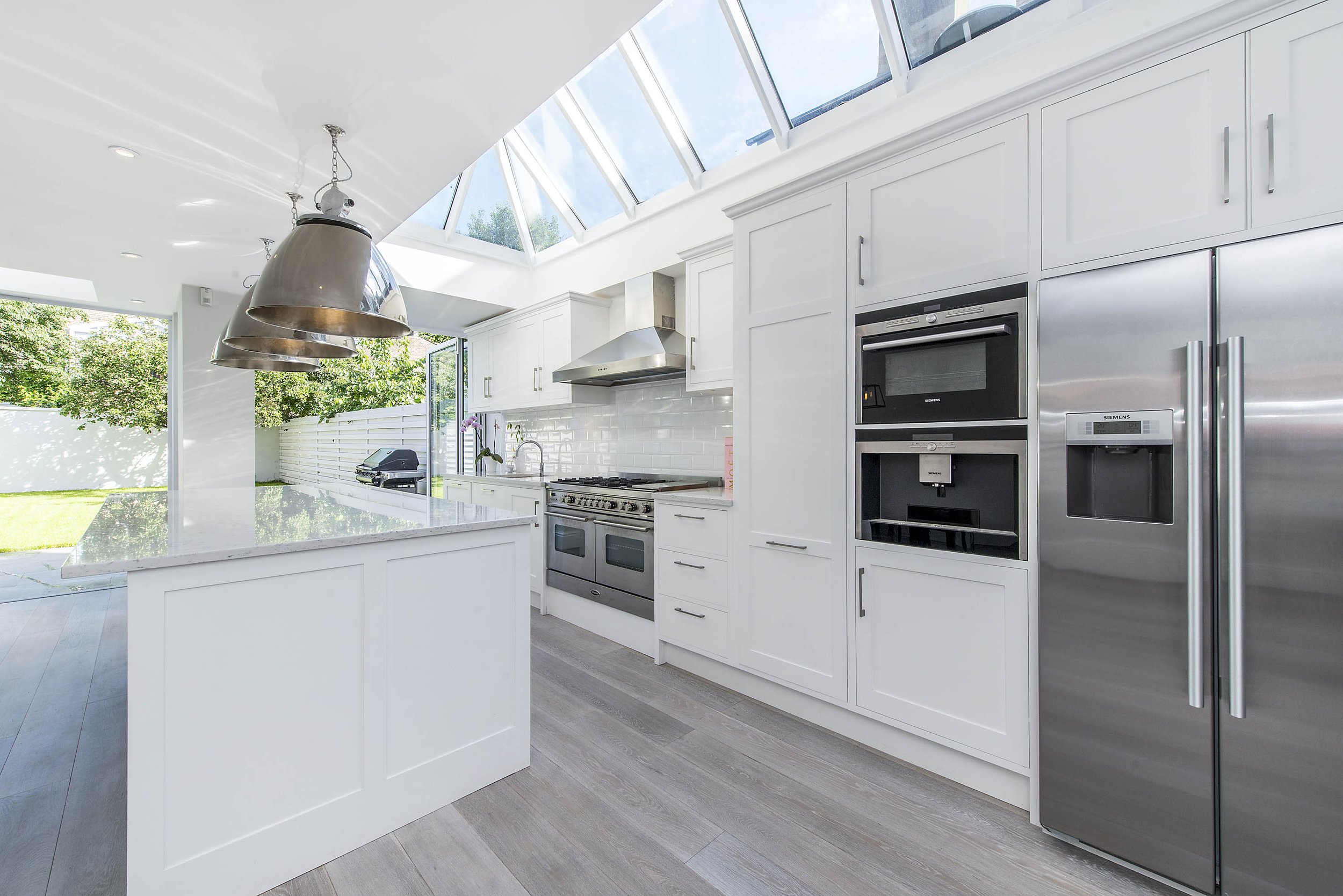 Bright and spacious modern kitchen with white cabinets, high-end appliances, and a stainless steel fridge. The dining table with dark chairs rests on elegant hardwood floors. Large windows offer a garden view, filling the room with natural light and