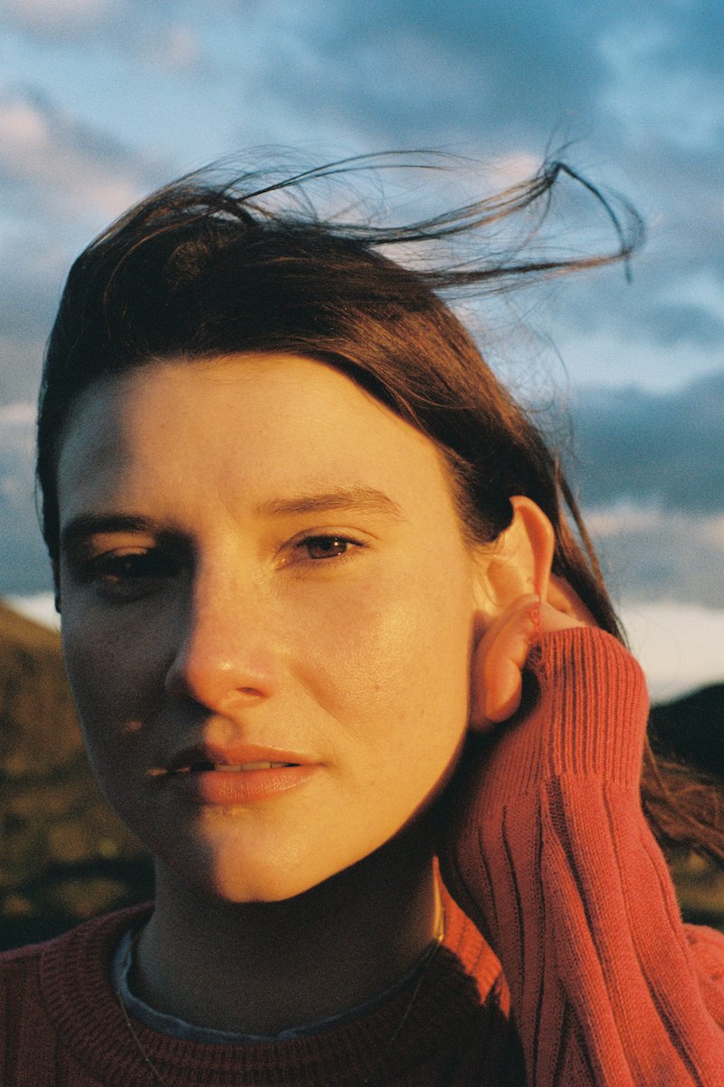 Close-up of a young woman with dark hair, slightly windblown, wearing a red sweater, outdoors during sunset with a cloudy sky in the background.