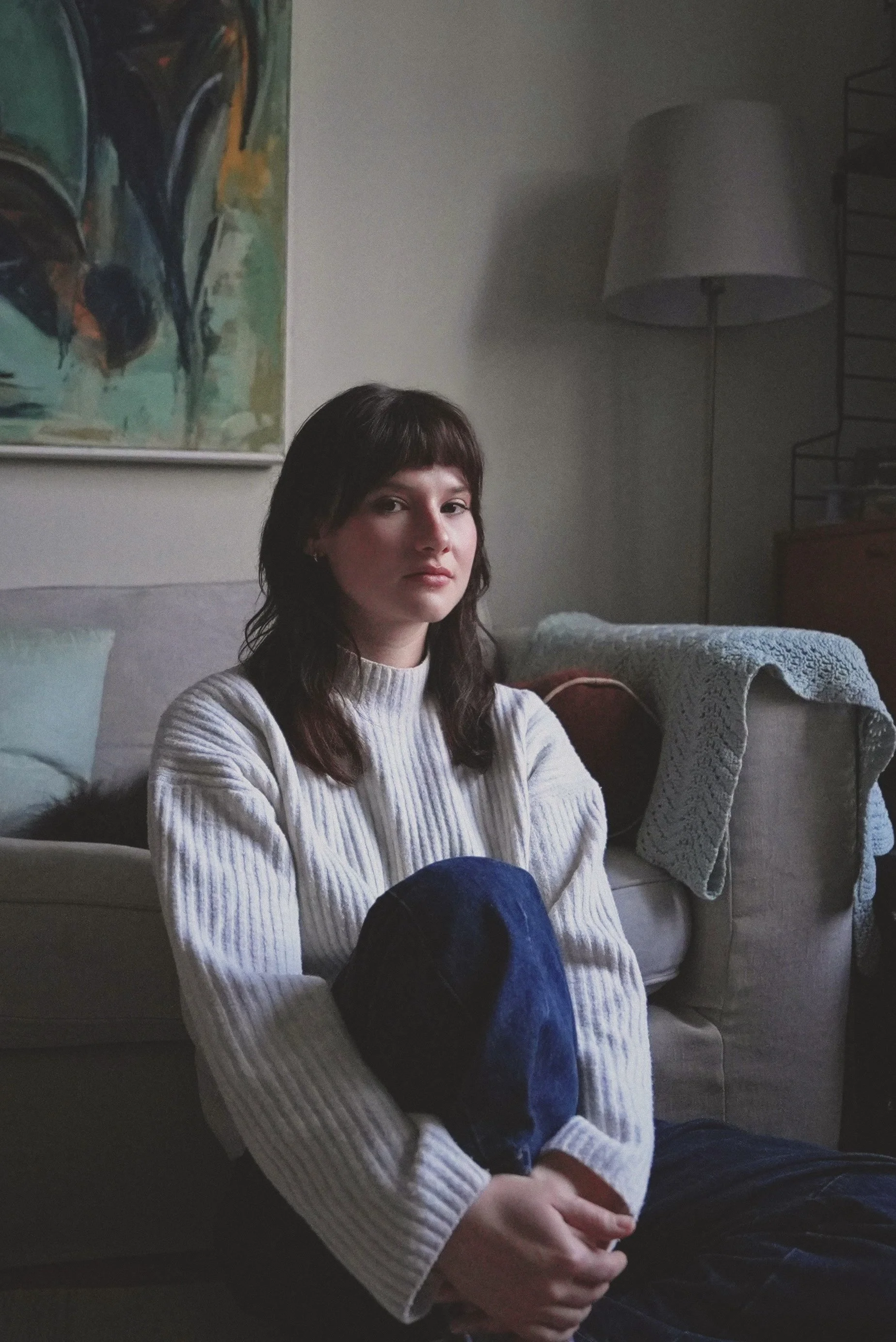 A young woman with dark brown hair, wearing a white knit sweater, sitting on a beige couch. She has a calm expression and is looking towards the camera. Behind her, a colorful abstract painting hangs on the wall. To her right, a large white lamp and a black metal shelf with decorative items are visible. A light blue pillow rests on the couch, and a light-colored blanket is draped over the back of the couch.
