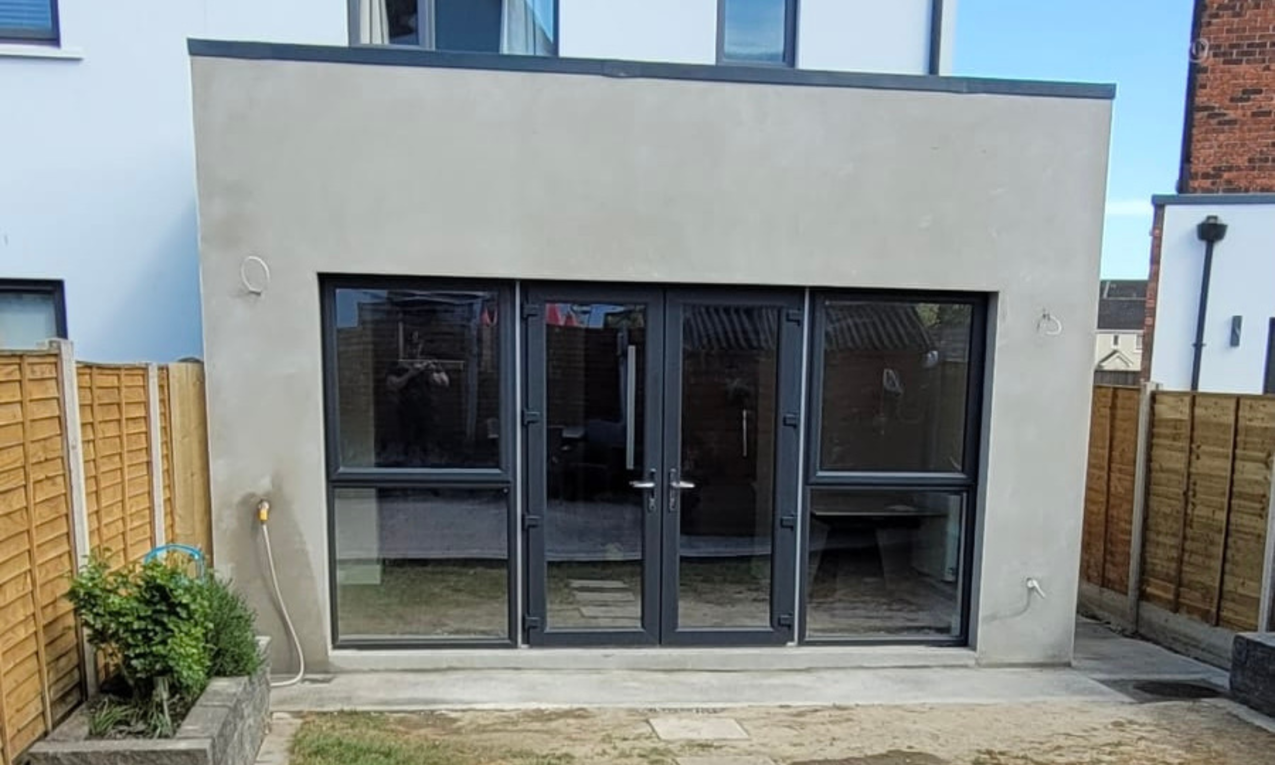 A modern residential back patio with a small yard, featuring a second-floor balcony, wooden fences on both sides, and a large glass door leading inside. There is a garden hose on the ground and some potted plants on the left.