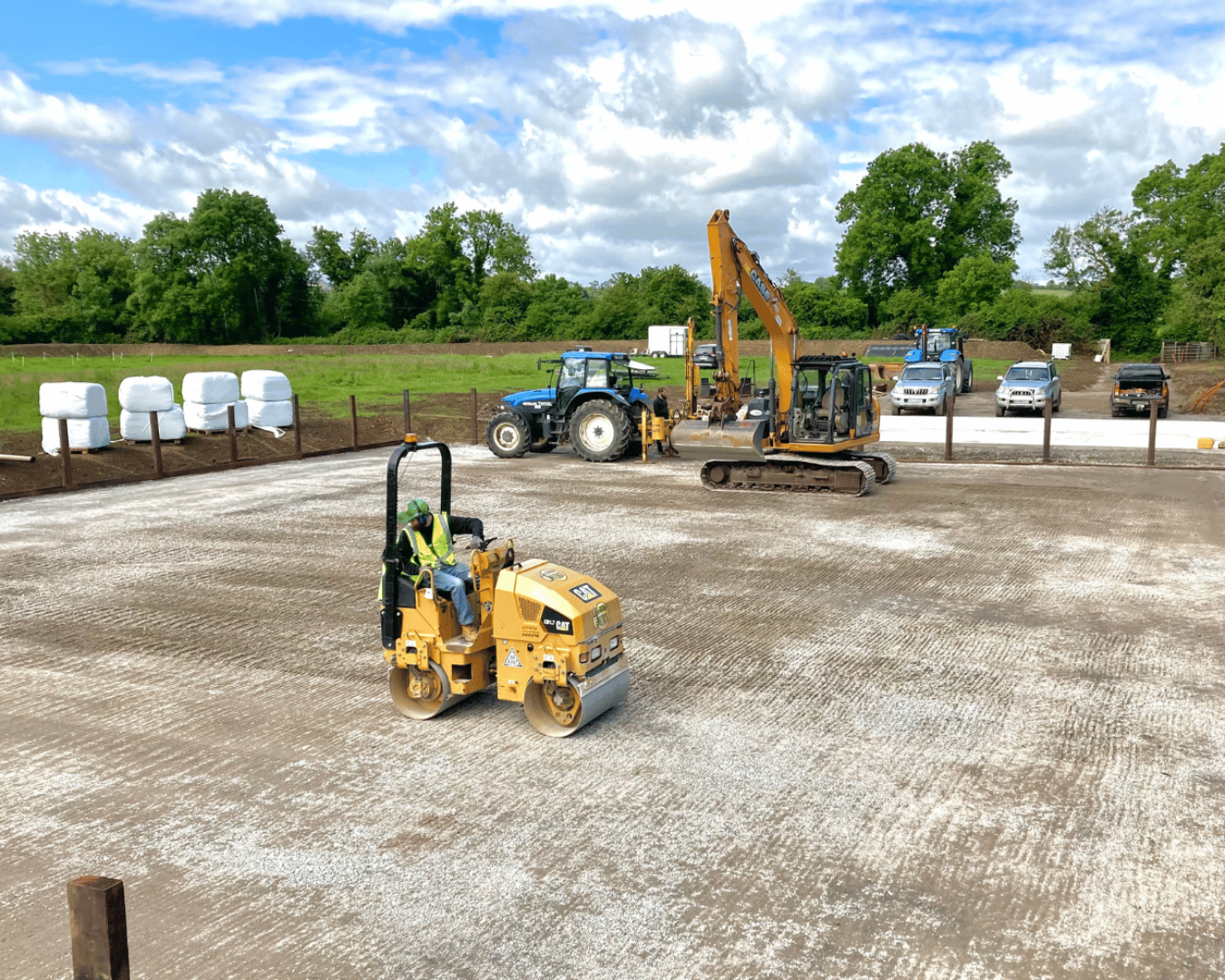 Construction site with a worker operating a compactor, a small excavator, a tractor, and several parked cars against a backdrop of green trees and a partly cloudy sky.