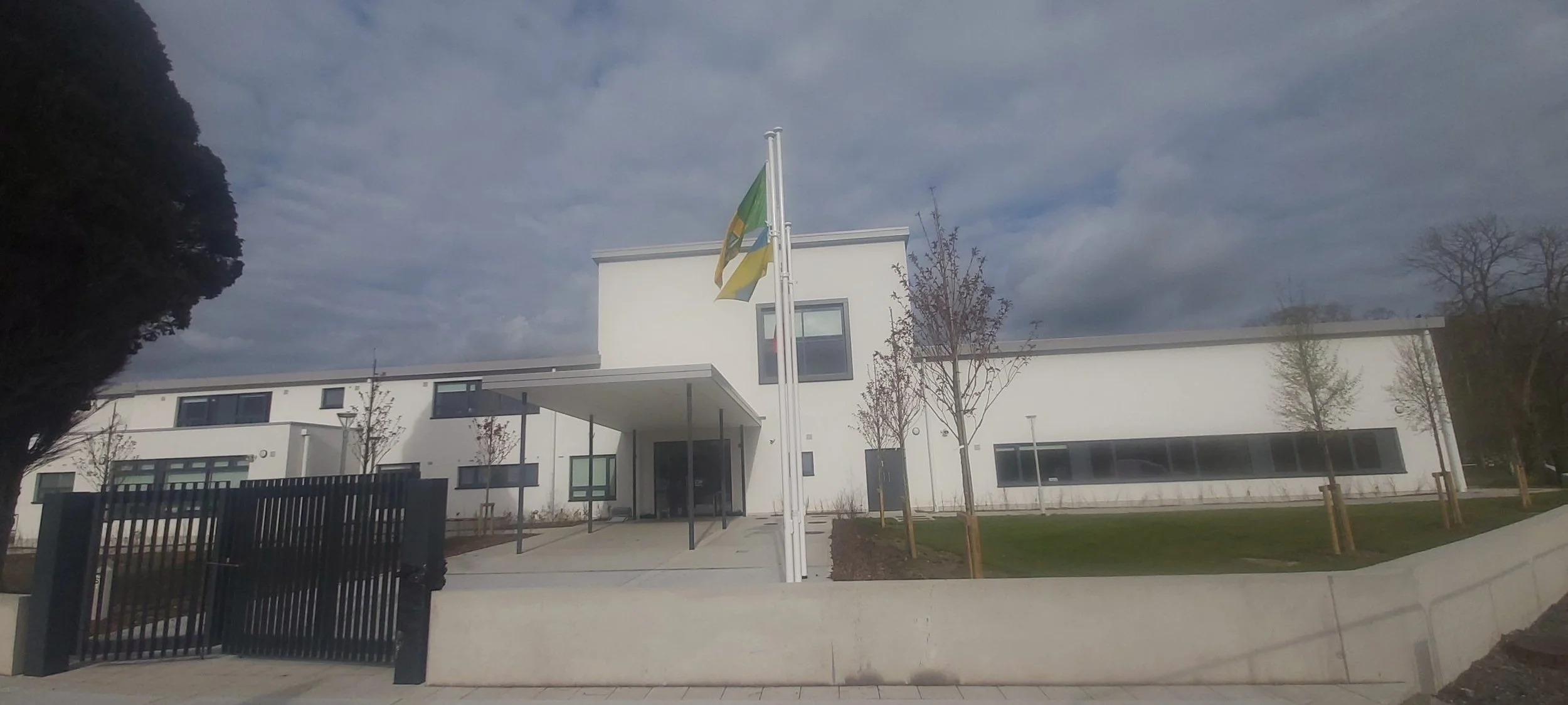 Modern white building with a flagpole displaying two flags, surrounded by young trees, a black fence, and a concrete sidewalk, under a cloudy sky.