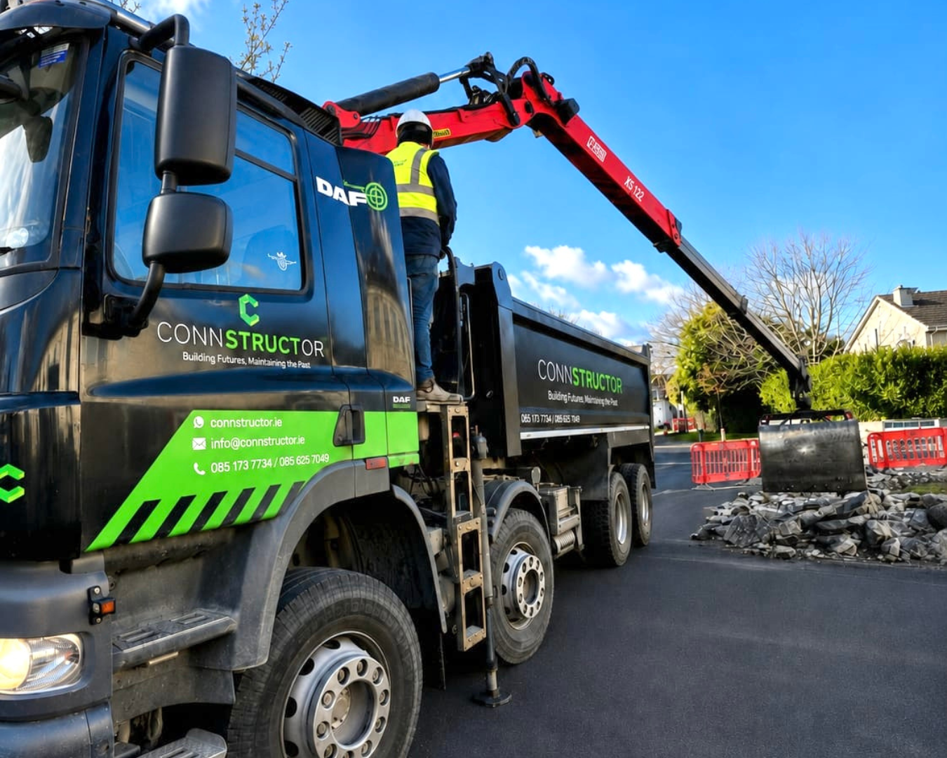 A construction truck with green and black markings and a red crane arm lifting debris and rocks on a street. A worker in safety gear is standing on the truck's side, and there are orange safety barriers in the background.