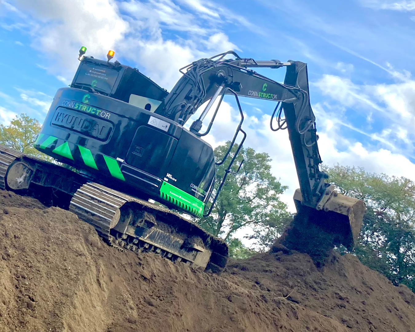A Hyundai excavator with the brand name visible on its side, working on a large dirt mound outdoors during daytime. The sky is partly cloudy, and there are trees in the background.