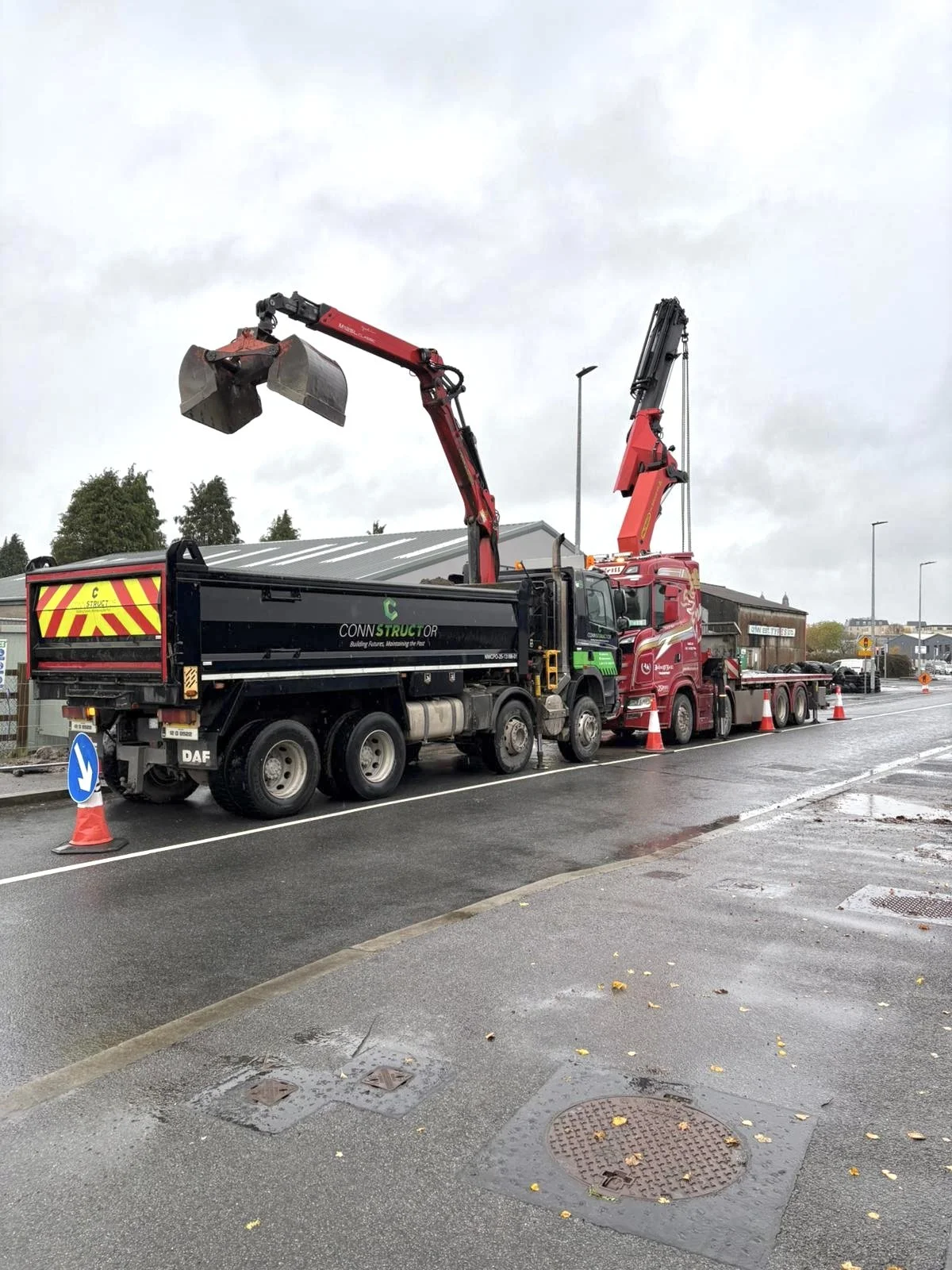 A large construction truck with a crane arm lifting a piece of heavy equipment on a wet road, surrounded by orange traffic cones and traffic signs, with overcast sky in the background.