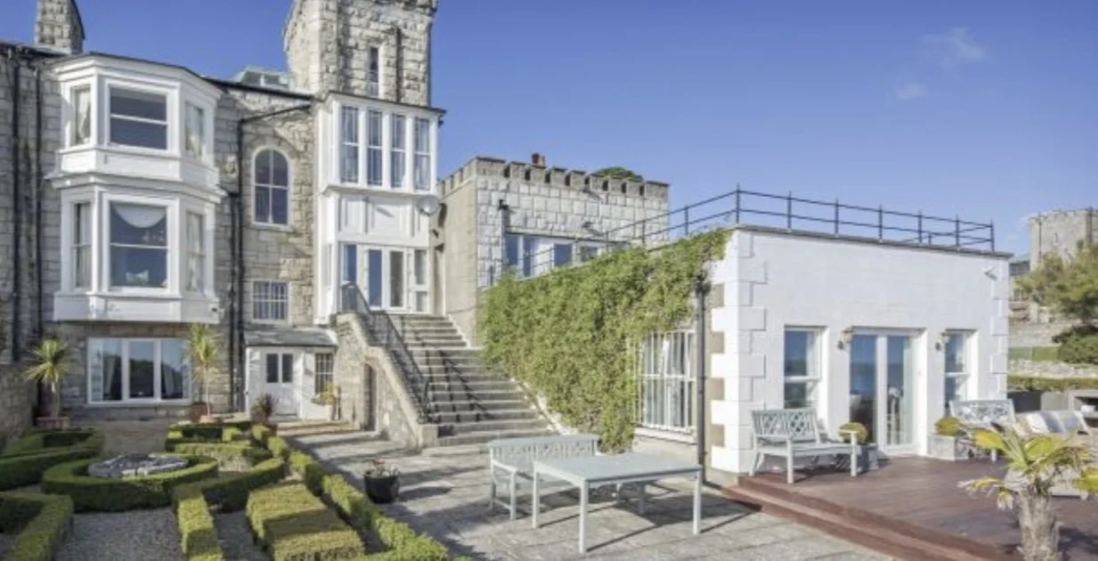 A large stone building with white bay windows, a staircase, and an outdoor area with benches, plants, and landscaped hedges.