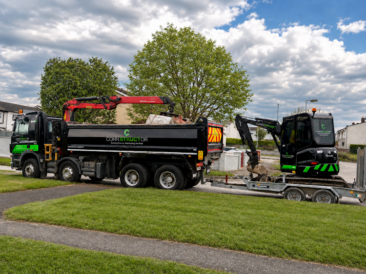 Construction trucks parked on a grassy area near sidewalks, with trees, buildings, and cloudy sky in the background.