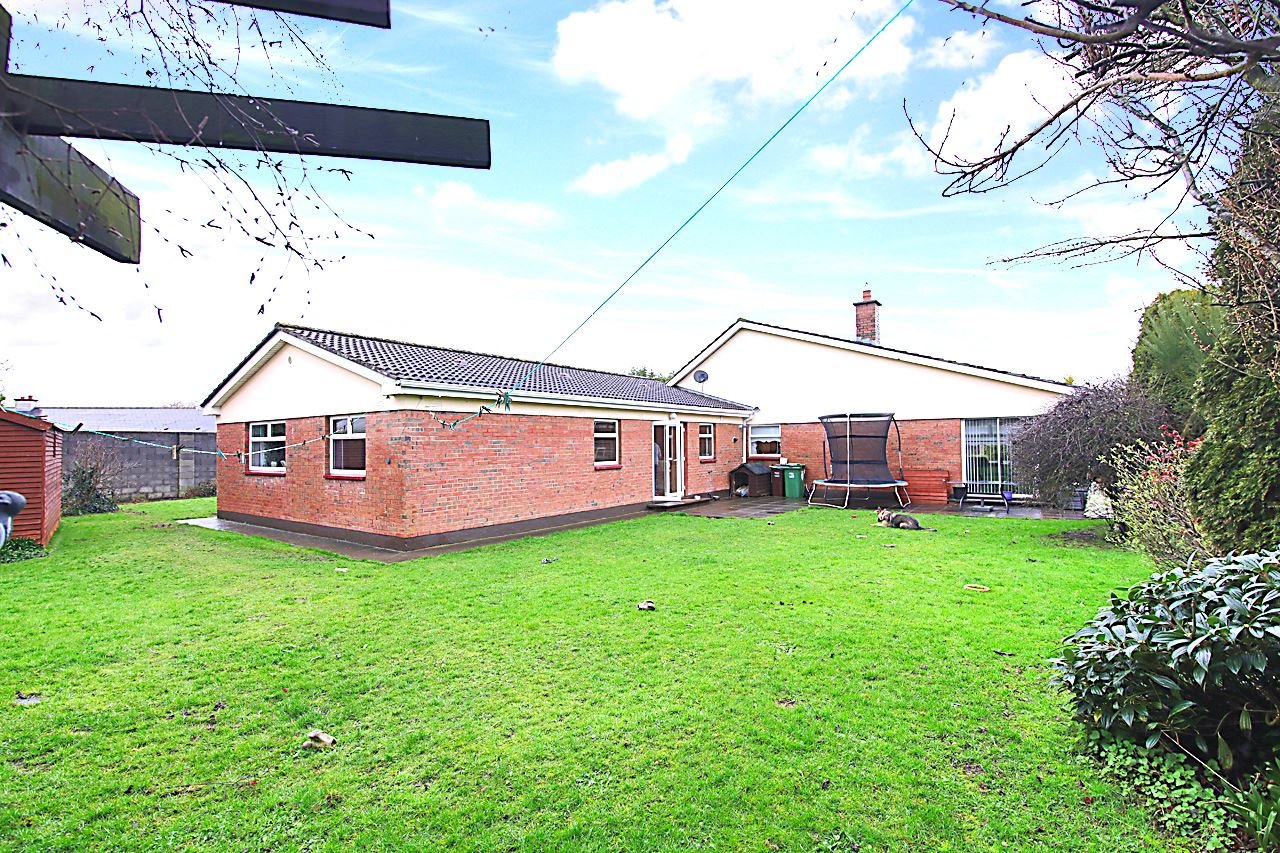 Backyard view of a house with brick walls, green lawn, trampoline, garden furniture, and trees, under a partly cloudy sky.