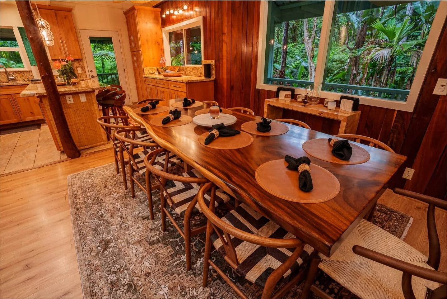 A dining room featuring a long, polished wooden table set with eight black cloth napkins. The room has large windows showcasing lush green jungle outside. The walls are made of wood paneling, and a small side table is decorated with glassware and fra