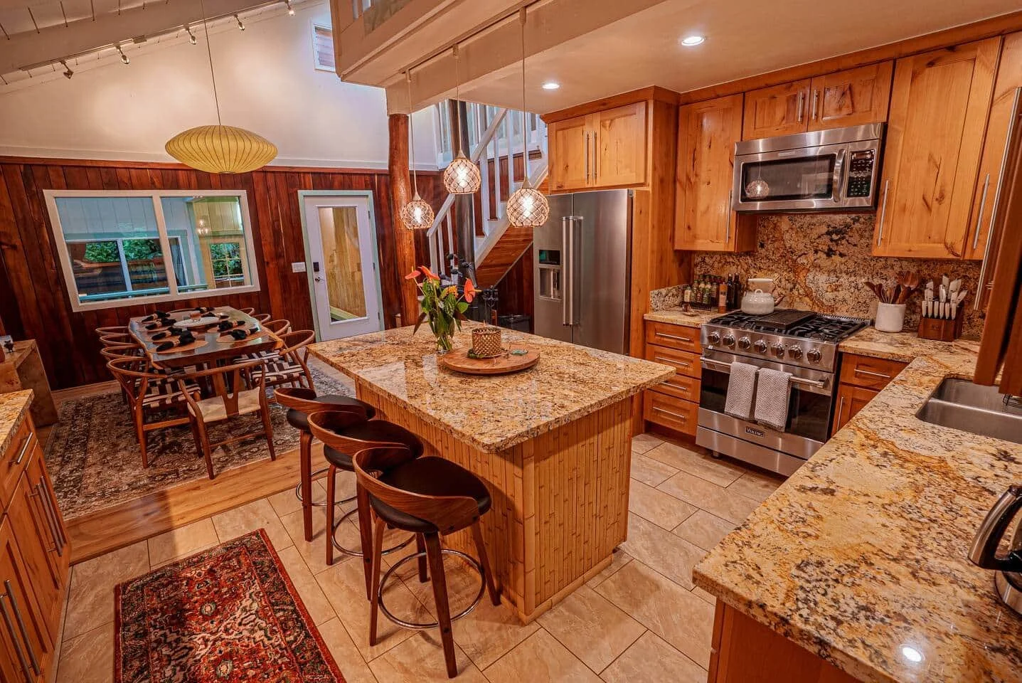 A kitchen with wooden cabinets, a granite countertop island, stainless steel appliances, a dining area with a wooden table and chairs, pendant and ceiling lights, and decorative items on the countertops.