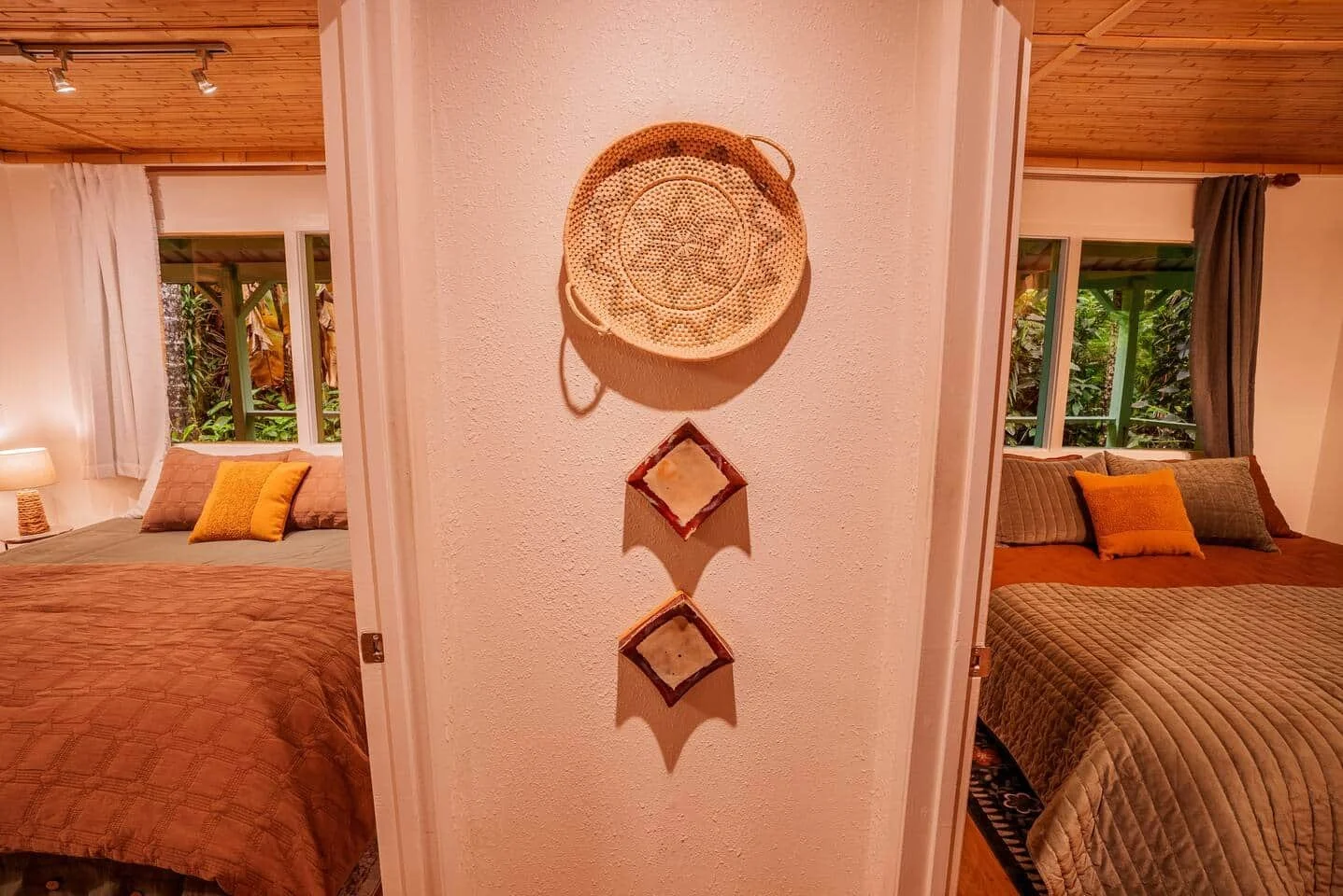 A bedroom with two beds, one on each side of the doorway, with pillows and blankets; lamps and windows with trees outside; a wall with a decorative round basket and two square wall hangings.