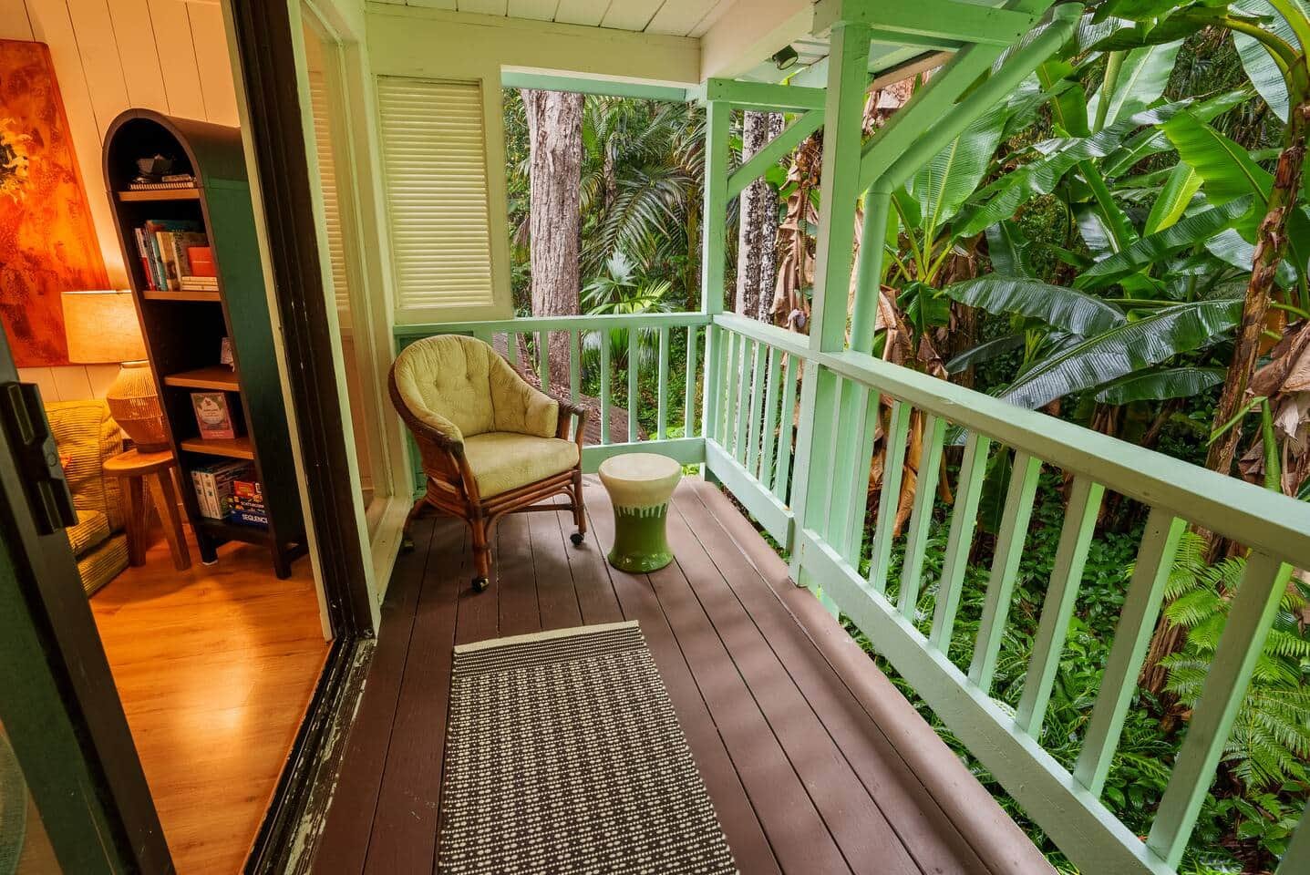View of a small balcony with wooden flooring and white railing, overlooking lush tropical greenery including large leaves and trees.