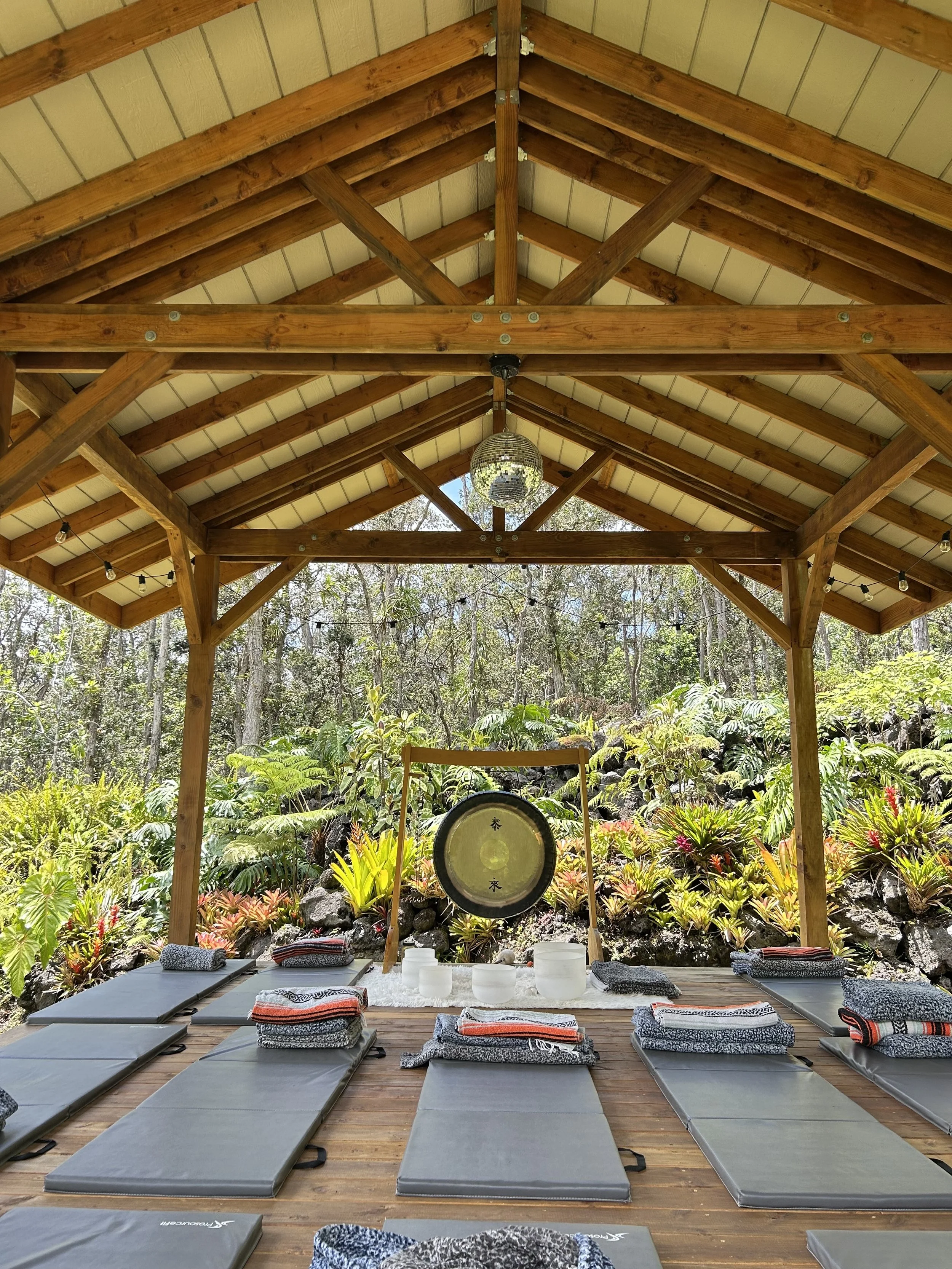 Outdoor yoga or meditation space under a wooden pavilion with mats, towels, and a gong, surrounded by lush greenery and trees.
