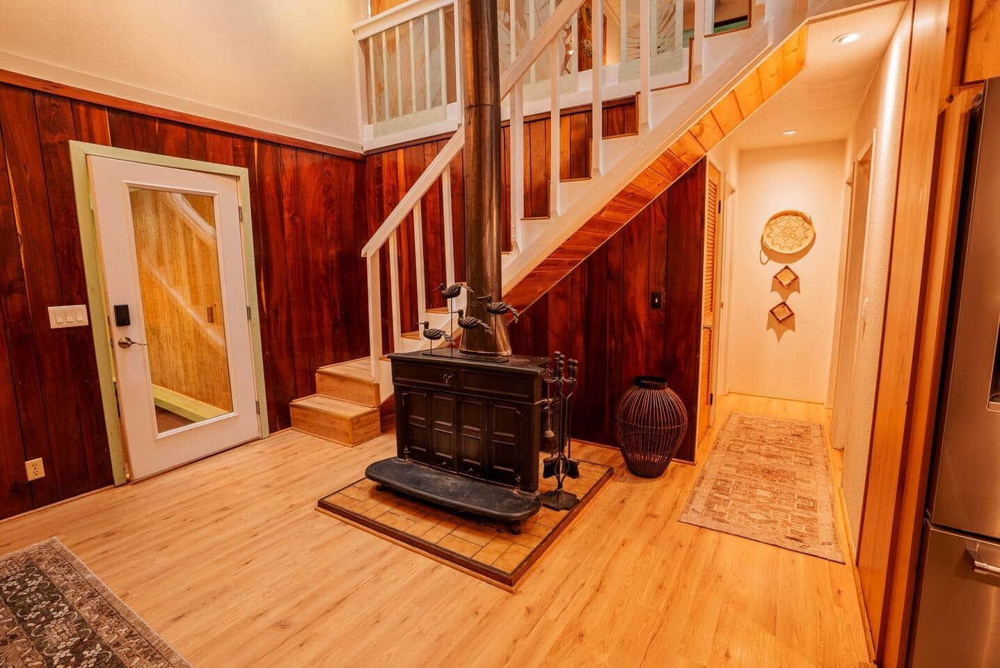 Interior view of a house with wooden walls and flooring, a staircase leading upstairs, and a wood stove on a tiled platform. There are decorative wall hangings and a woven basket near the hallway.
