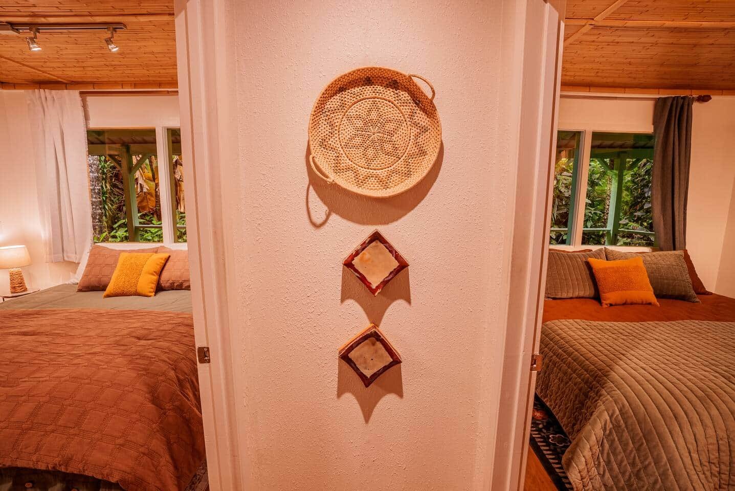 Interior view of a bedroom with two beds separated by a wall with decorative objects hanging on it, including a woven basket and two small square plates, with windows showing greenery outside.