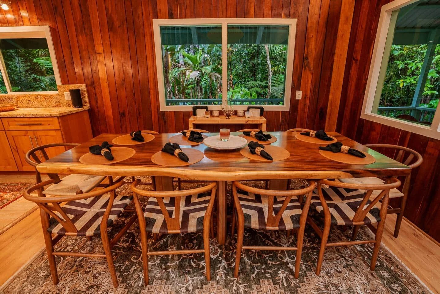 A rustic wooden dining room with a large oval table set with black napkins and brown placemats, a central candle, overlooking lush green jungle through three windows.