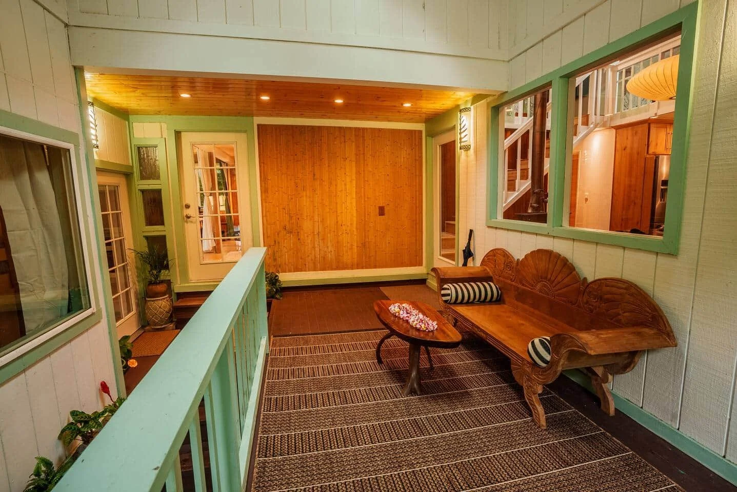 Interior porch with a wooden bench and coffee table, striped pillows, potted plants, and windows revealing a staircase and wood-paneled walls.