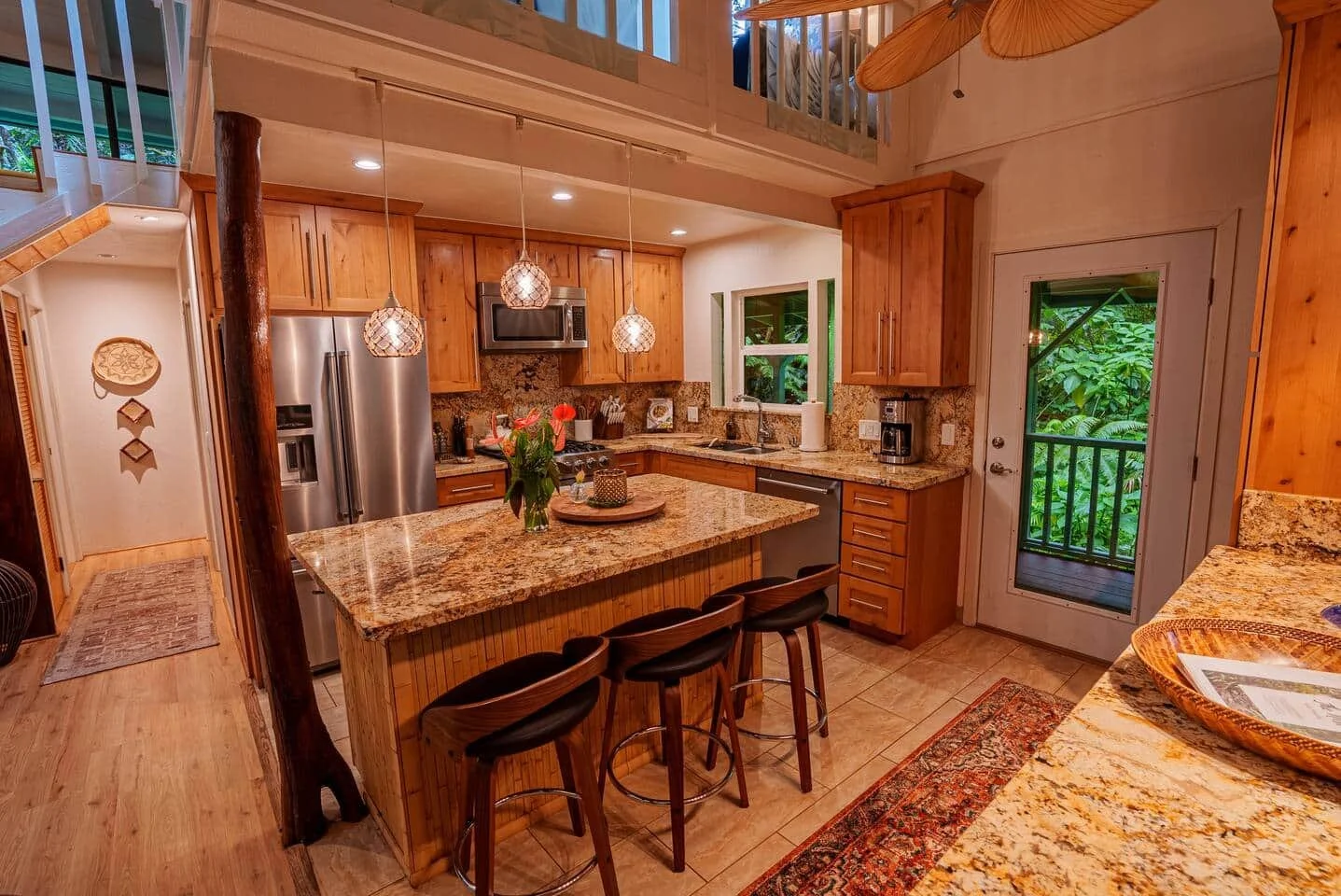 A kitchen with wooden cabinets, granite countertops, and a central island with three chairs. Stainless steel refrigerator, microwave, and coffee maker. Pendant lights hang above the island, and there is a window showing green foliage outside.