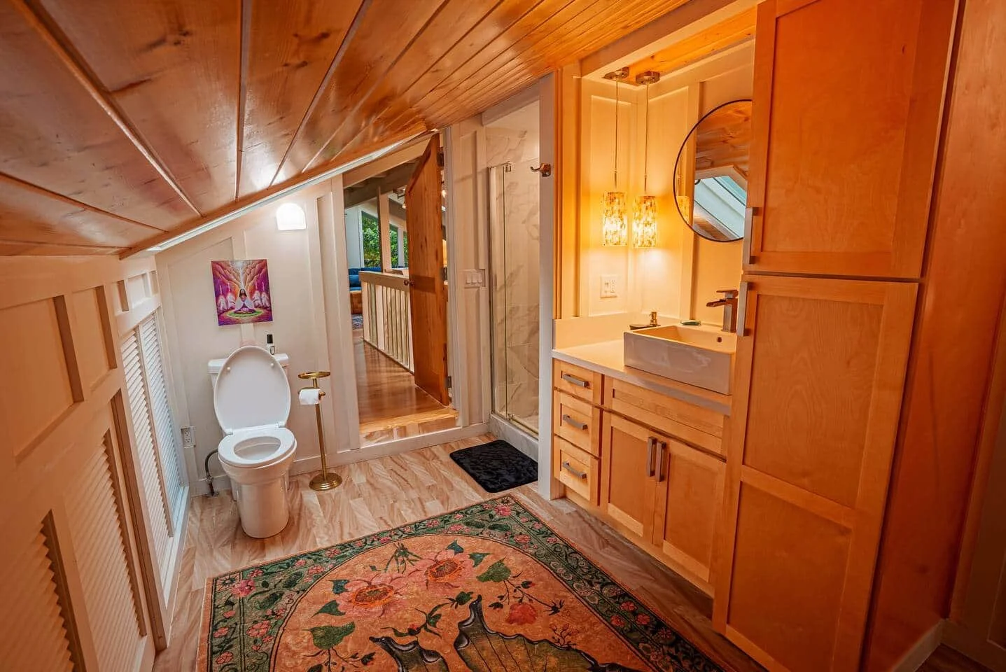 Small rustic bathroom with sloped wooden ceiling, white toilet, wooden cabinetry, a small sink with a round mirror, warm lighting, and a colorful rug on wooden floor. Visible hallway and living room in background.
