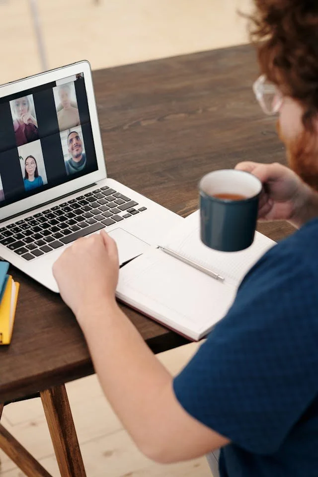A person with curly hair and glasses holding a coffee mug meets with four other colleagues on a video call on a laptop, with a notebook and pen on the table.