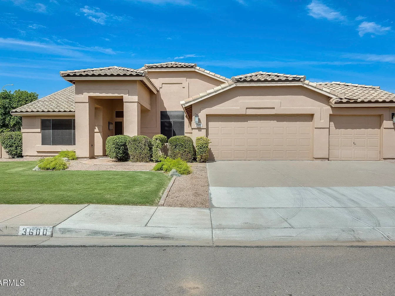 A modern, beige, single-family house with a tiled roof, front yard with green lawn, bushes, and plants, two-car garage, and a sidewalk in a sunny neighborhood.