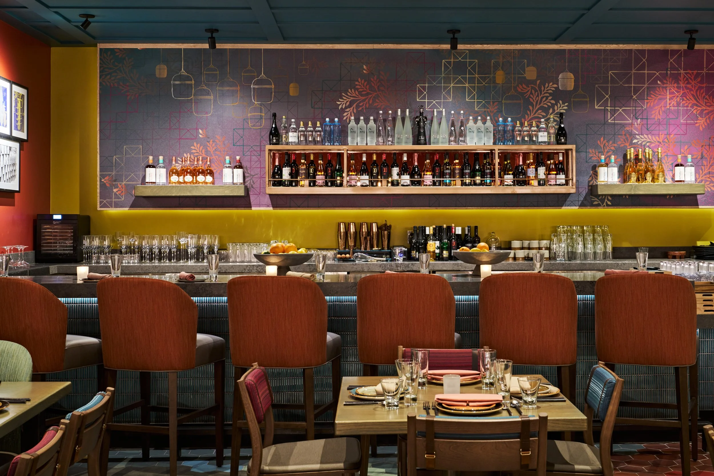 View of a stylish bar area with a colorful wall, bottles of alcohol on shelves, and a neatly set dining table with glasses and utensils.