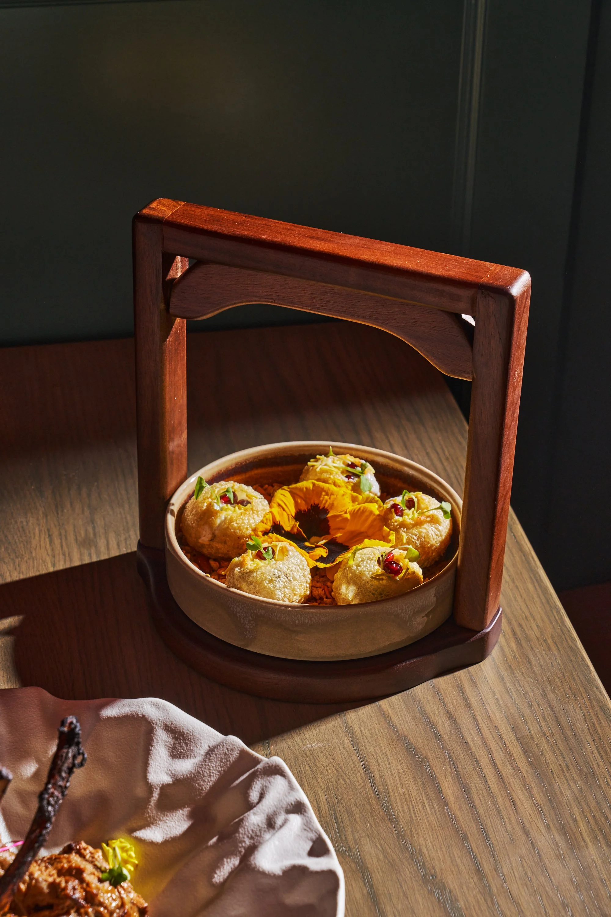 A bowl of round, cream-colored appetizers garnished with small green herbs and red berries, placed inside a wooden tray with a decorative handle, on a wooden table with a large yellow flower in the center.