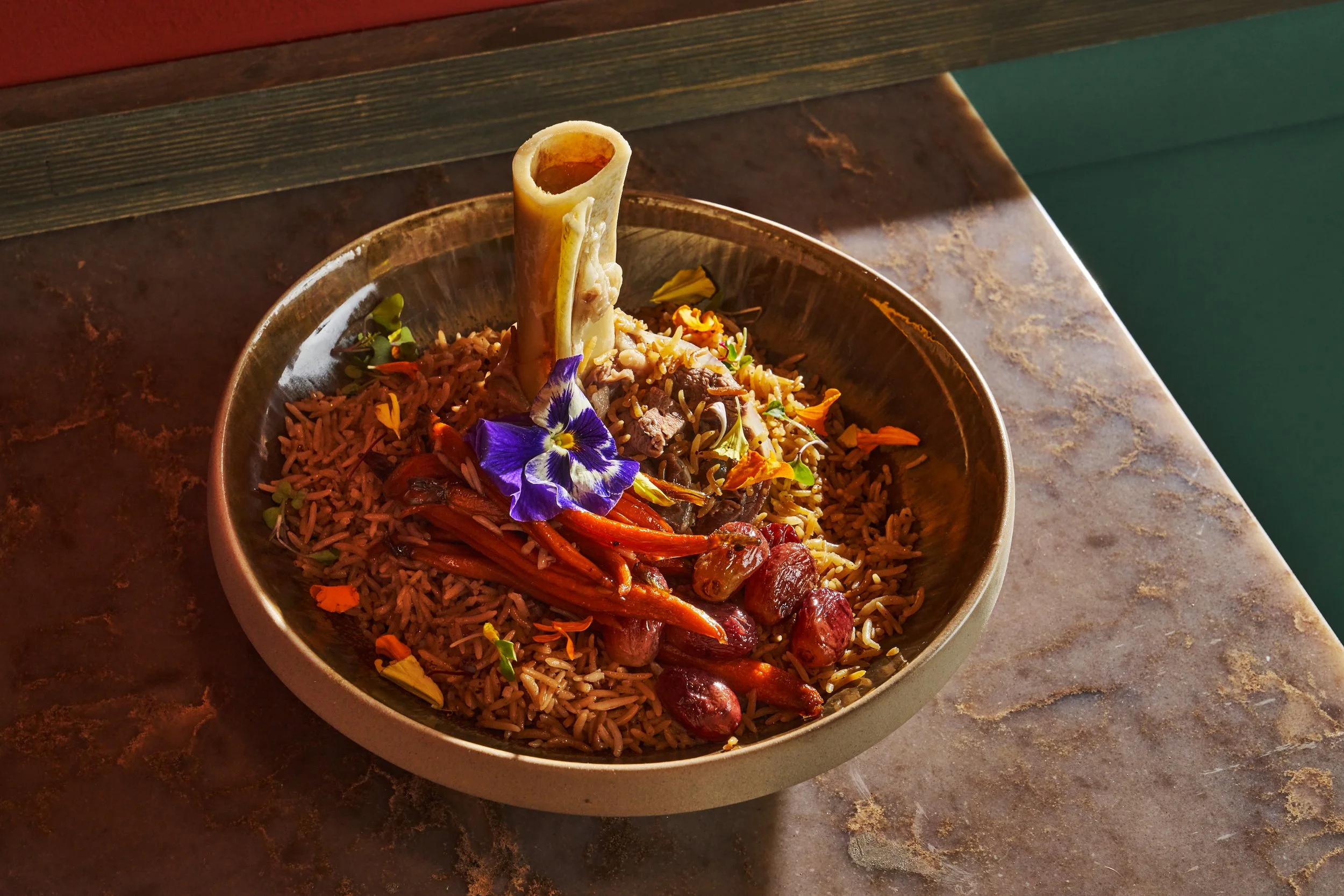 A bowl of cooked rice with vegetables, garnished with an edible purple flower, served on a rustic brown table.