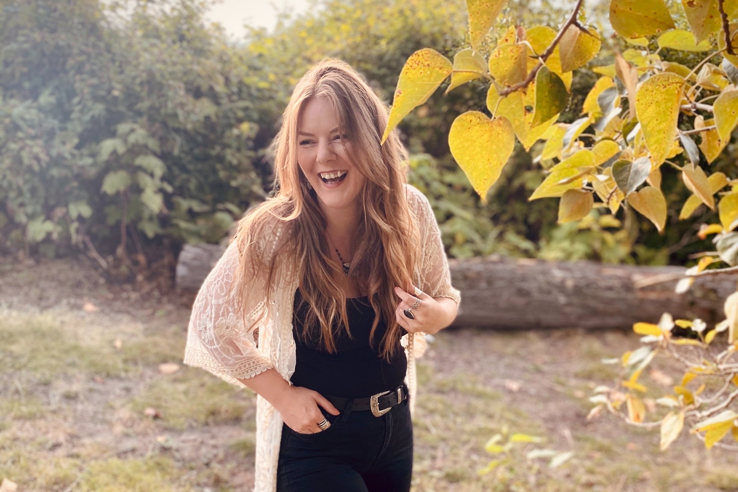 Woman with long hair smiling outdoors near green foliage and fallen leaves.