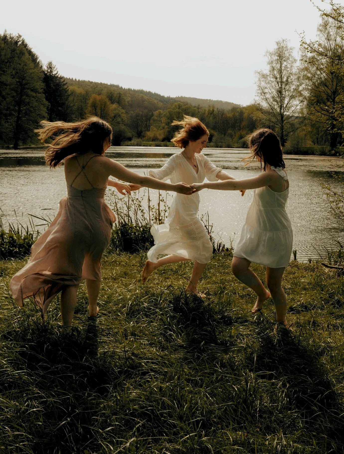 Three women in light dresses dancing in a circle on grass near a lake with surrounding trees.