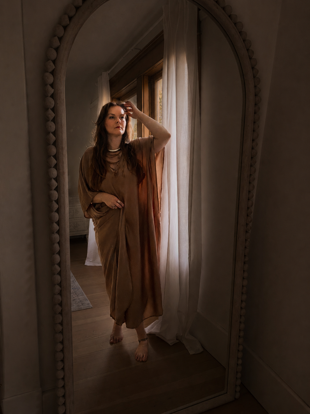 A woman in a brown flowing dress standing barefoot in front of a mirror, looking at herself and touching her hair, near a window with white curtains letting in natural light.