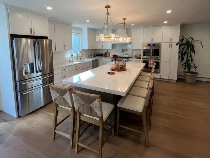 Modern kitchen with a large white island counter, surrounded by beige chairs, stainless steel appliances, white cabinets, pendant lights above, and a potted plant in the corner.