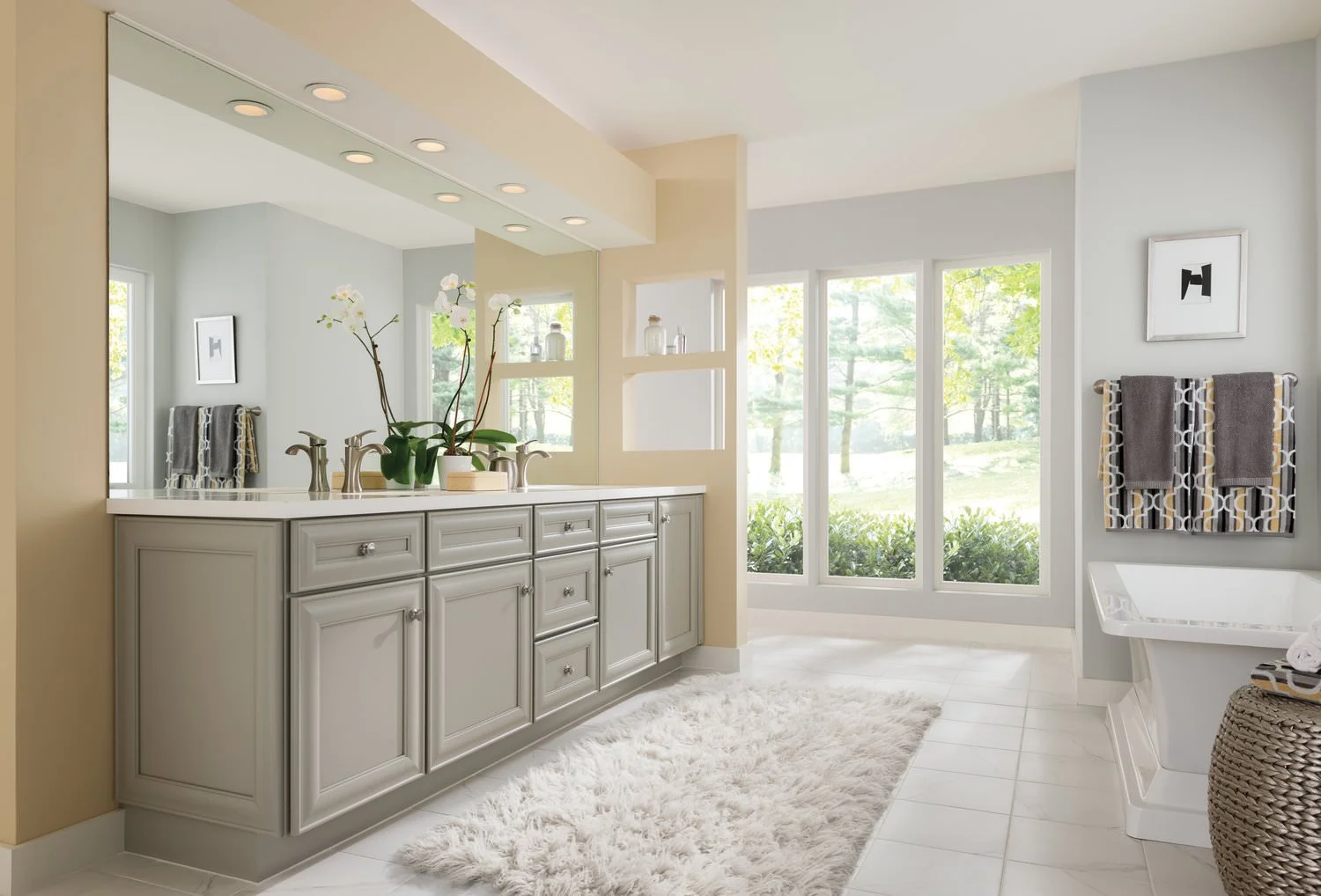 A bathroom with a large mirror, double sinks, and a window view of trees. There are towels on a rack, a framed picture on the wall, and a white rug on the tiled floor.