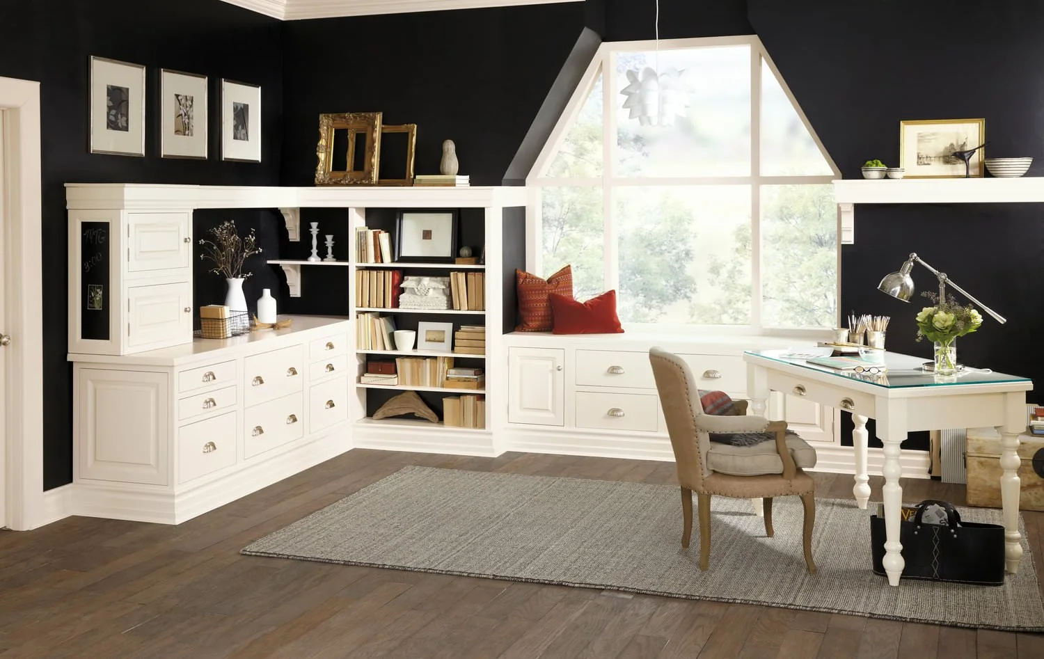 A home office with black walls, white built-in shelves and cabinets, a window seat with cushions, a white desk with a lamp and flowers, and a beige upholstered chair on a rug.
