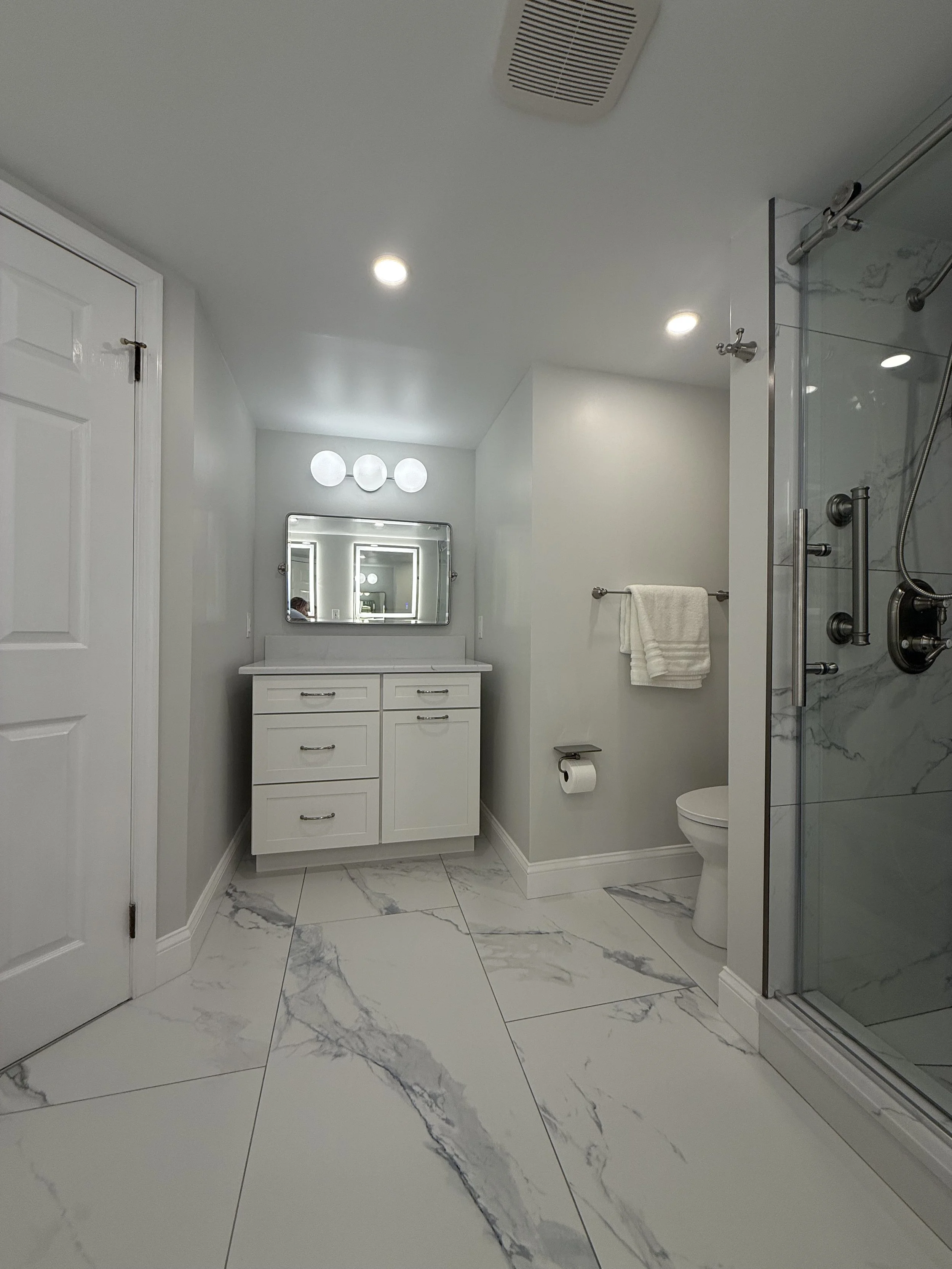 Modern bathroom with white marble tile floor, white vanity with mirror, toilet, towel rack with white towel, and glass shower with marble walls.