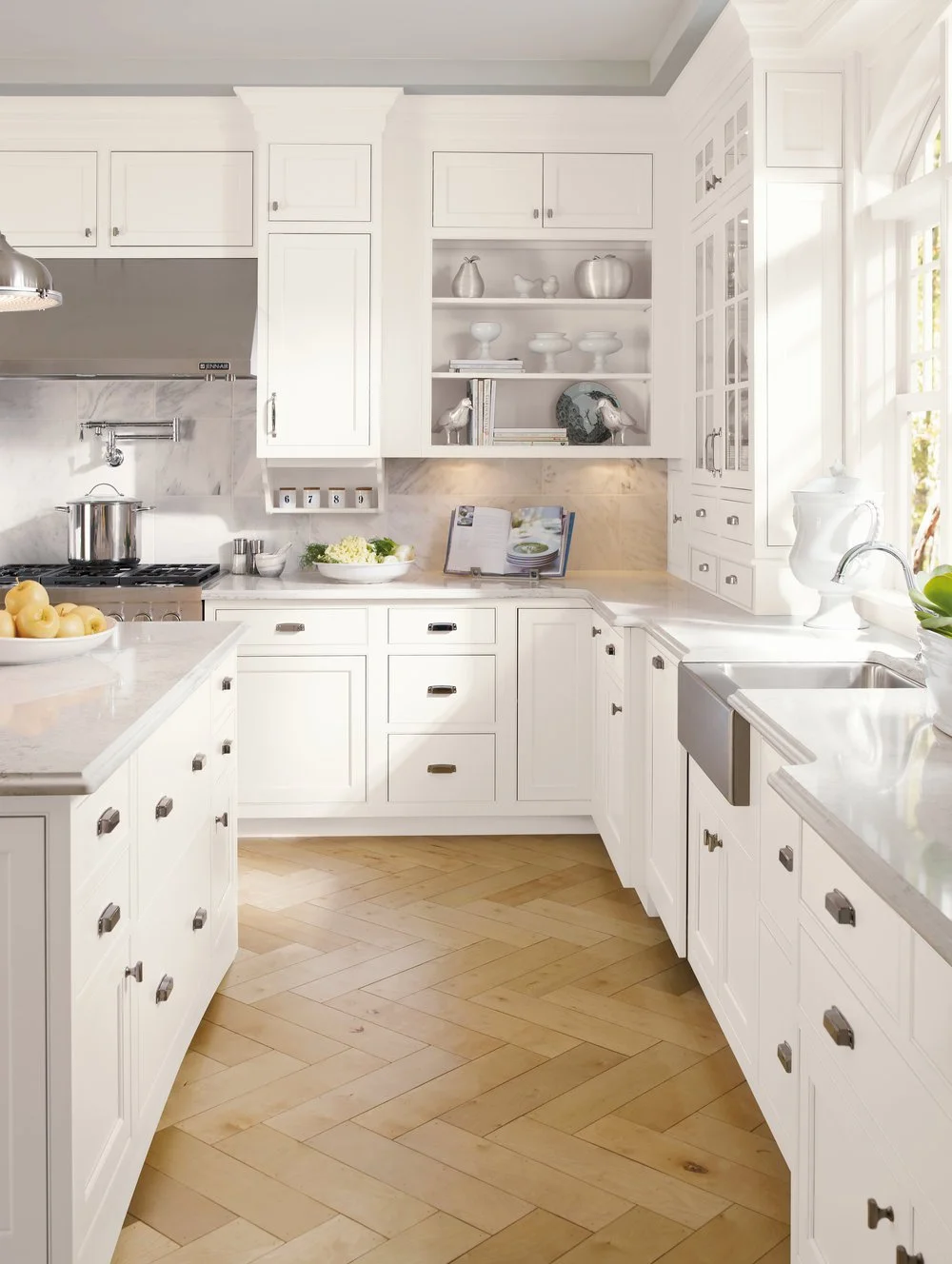 Bright white kitchen with marble countertops, open shelving with white and silver decor, and wooden herringbone floor.