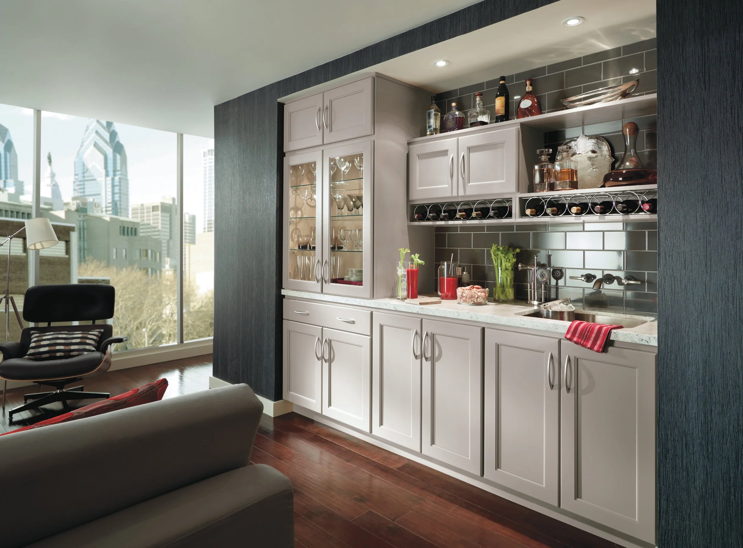 Modern kitchen with white cabinets, dark gray backsplash, and open shelving with bottles, glassware, and plants, next to living room with large windows and city skyline view.
