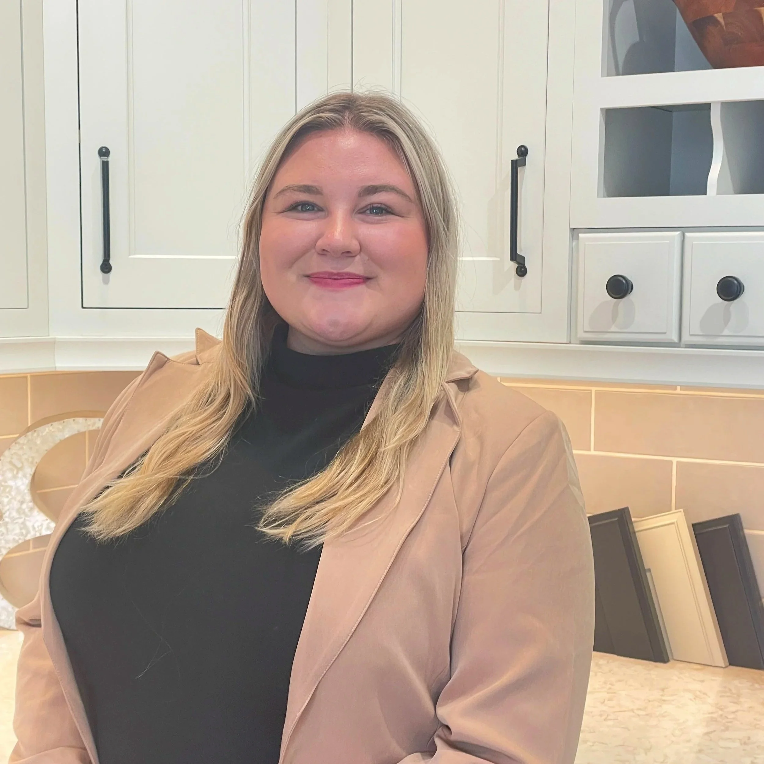 A woman with long blonde hair wearing a black turtleneck and a tan blazer smiling in a kitchen with white cabinets and black handles.