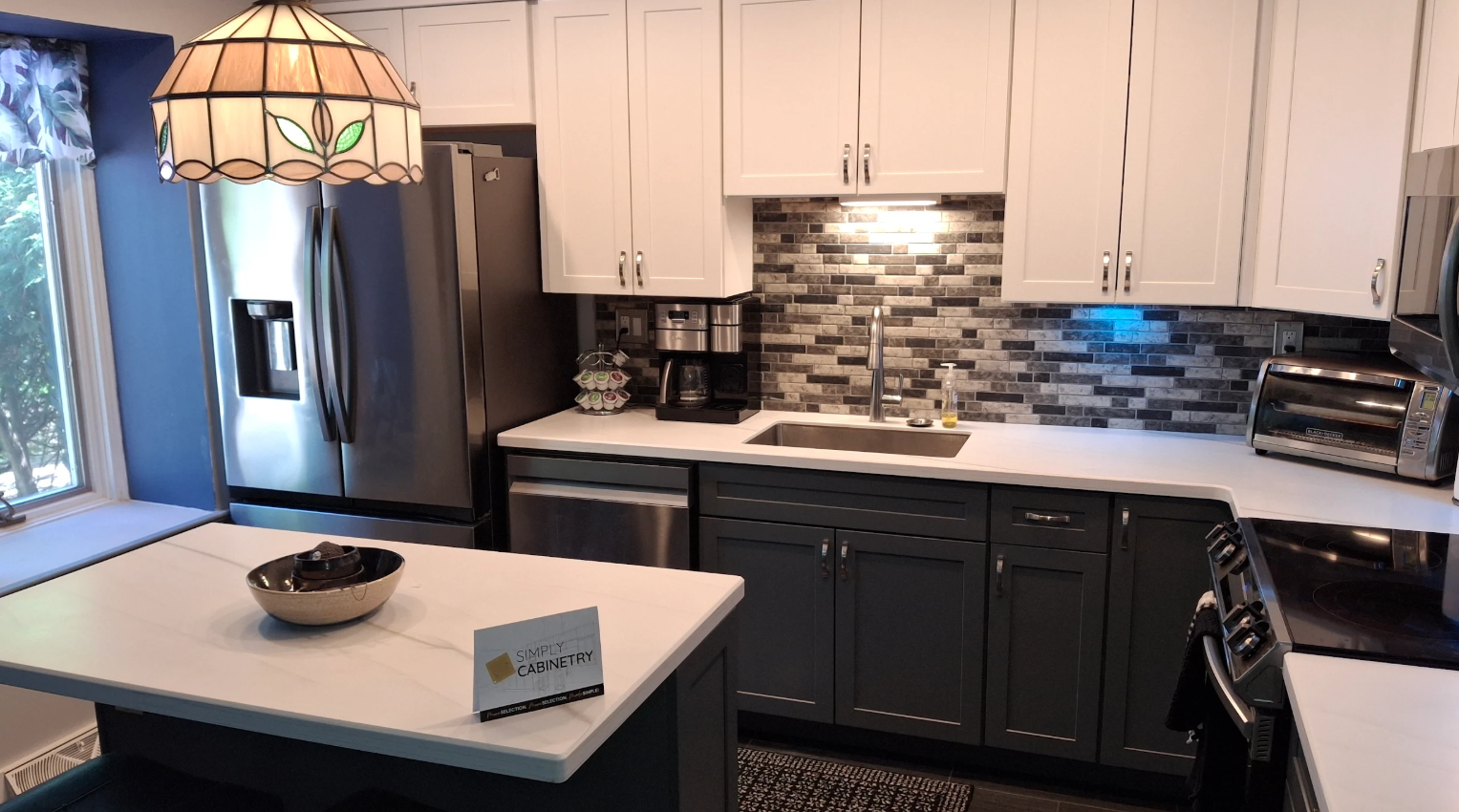 Modern kitchen with white upper cabinets and dark lower cabinets, stainless steel appliances, gray brick backsplash, and a counter with a coffee maker and toaster. A window with floral curtains is visible on the left.