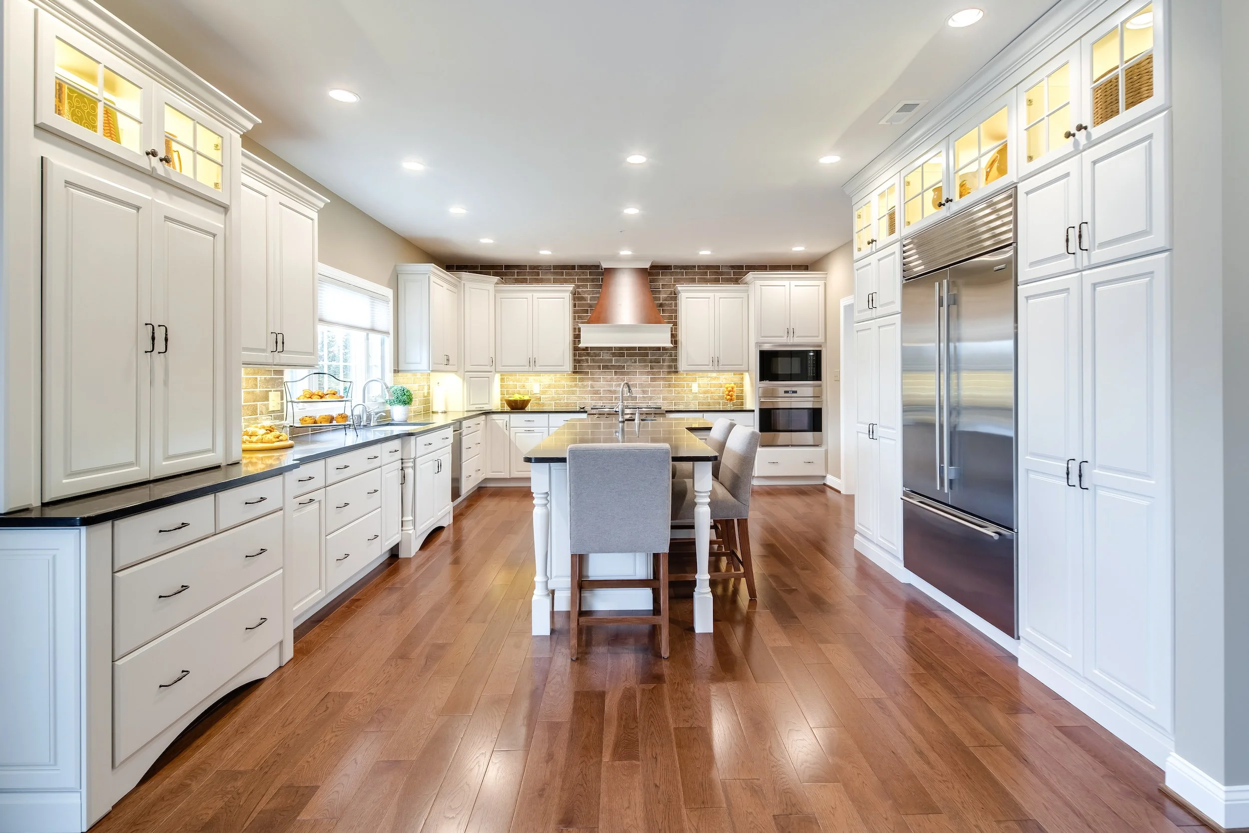 Modern kitchen with white cabinets, wood flooring, black countertops, a kitchen island with chairs, stainless steel appliances, brick back wall behind stove, and bright lighting.