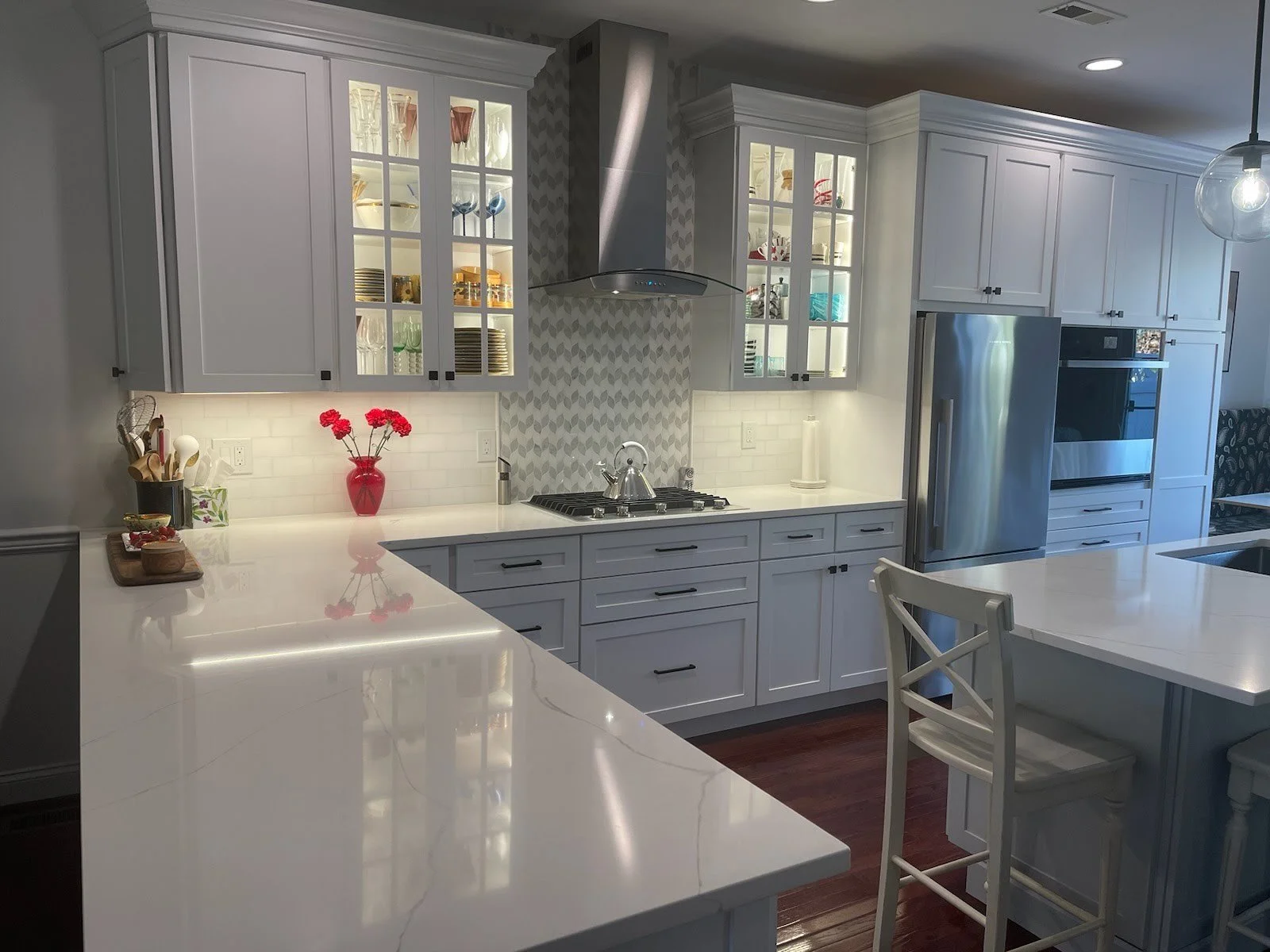Modern white kitchen with glass-front cabinets, stainless steel refrigerator, and a marble island. There is a red vase with flowers on the counter, a stove with a silver kettle, and a pendant light hanging from the ceiling.