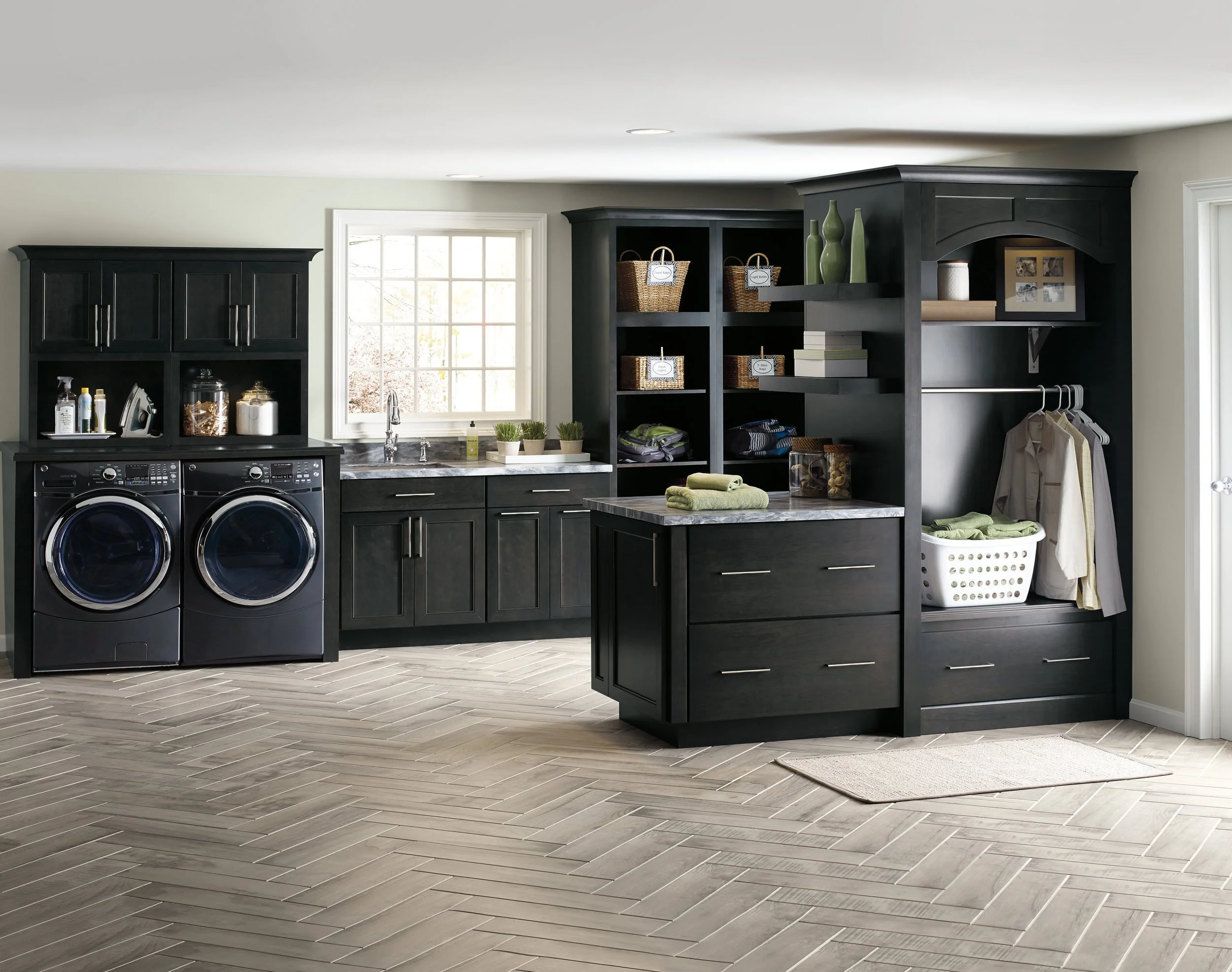 Laundry room with black cabinets, front-loading washer and dryer, window, countertop, open shelving with baskets and decor, and a laundry basket, with a chevron-patterned wooden floor.