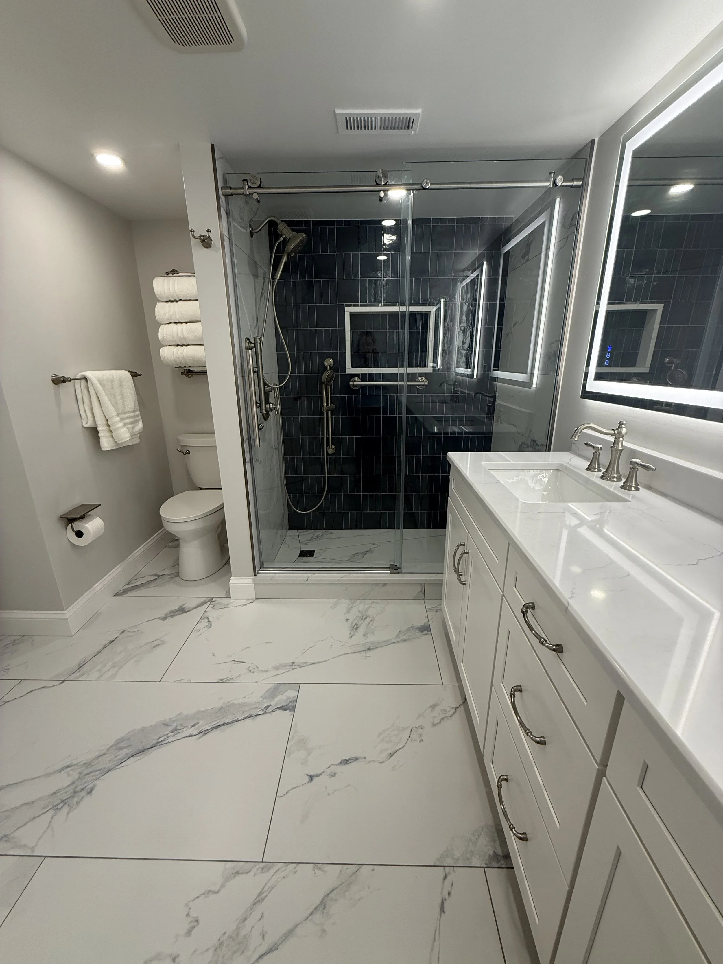 Modern bathroom with white marble floor tiles, a glass-enclosed shower with black tiles, a white vanity with a marble countertop, and a toilet with towel racks and shelves.