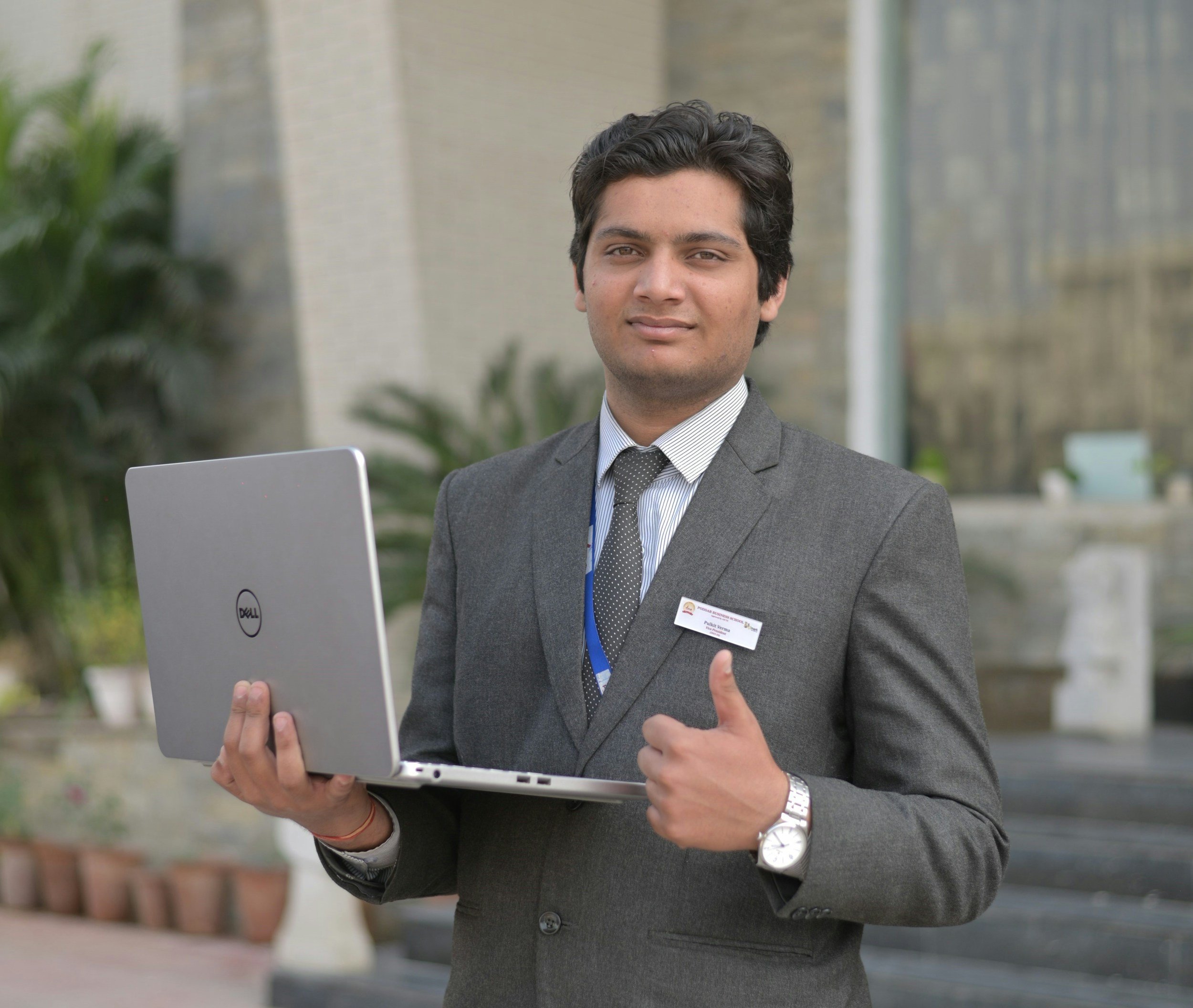Man in gray suit holding a laptop, giving a thumbs up, outdoors with greenery and buildings in background.