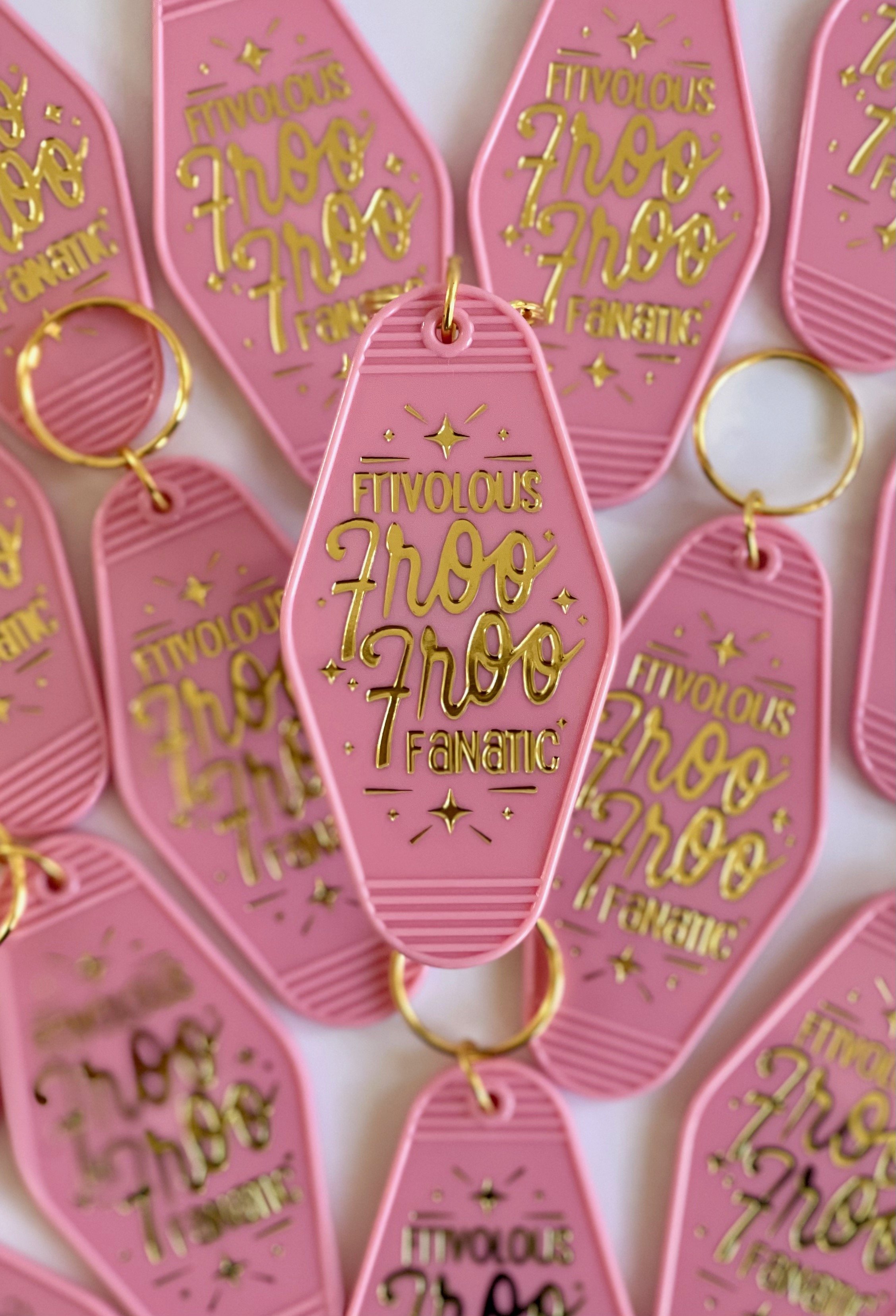A pink retro motel keychain featuring a design by KJ in gold foiling that reads, Frivolous Froo Froo Fanatic, and is adorned with gold foiled sparkles.