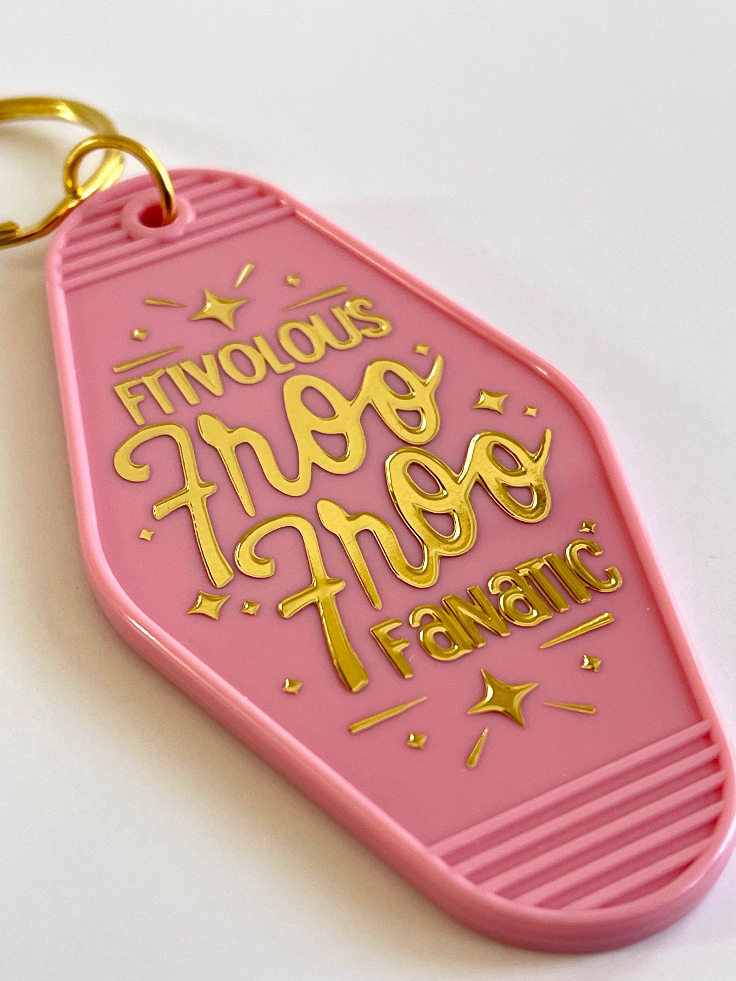 Details of a pink retro motel keychain featuring a design by KJ in gold foiling that reads, Frivolous Froo Froo Fanatic, and is adorned with gold foiled sparkles.