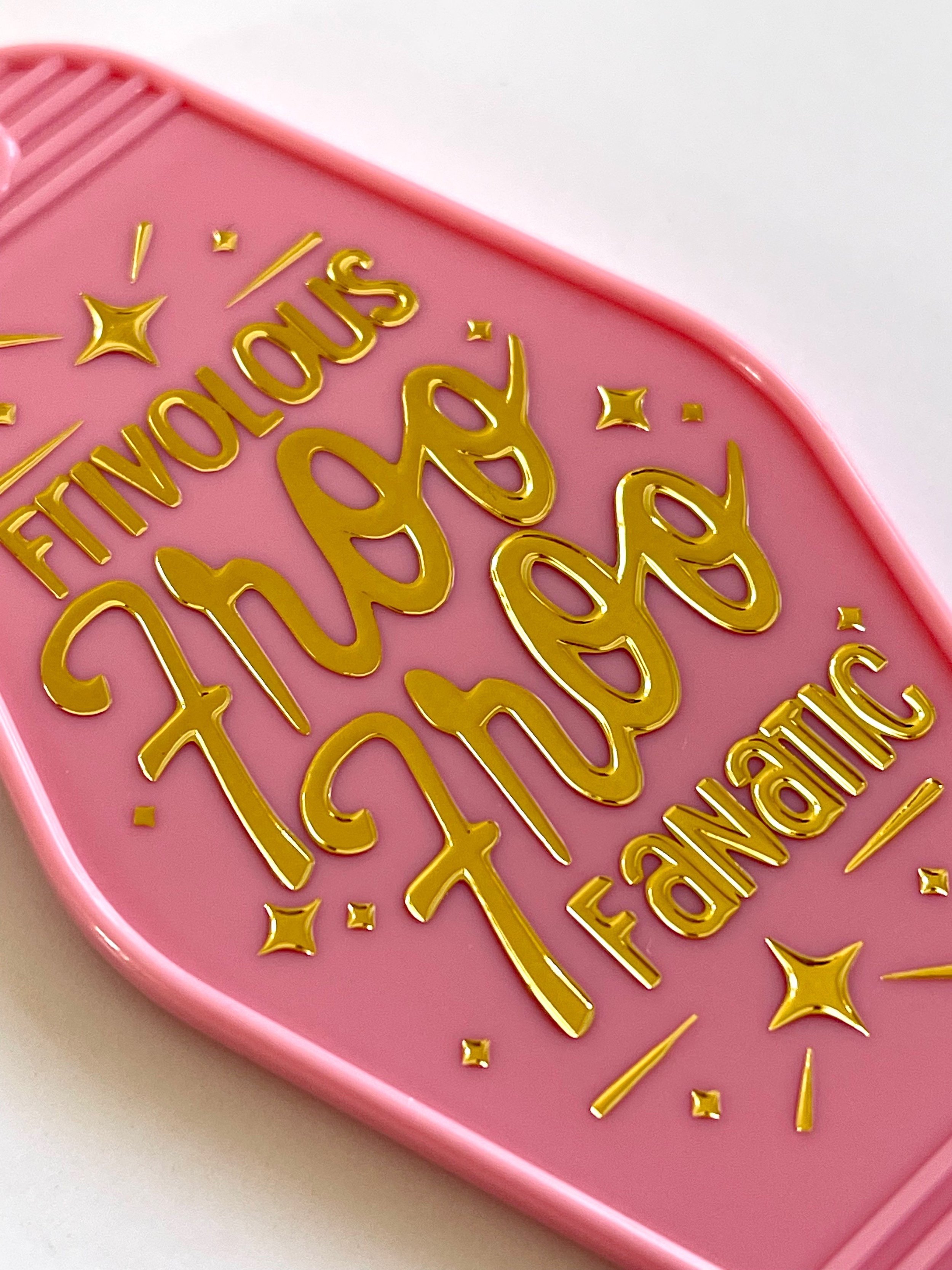 Details of a pink retro motel keychain featuring a design by KJ in gold foiling that reads, Frivolous Froo Froo Fanatic, and is adorned with gold foiled sparkles.